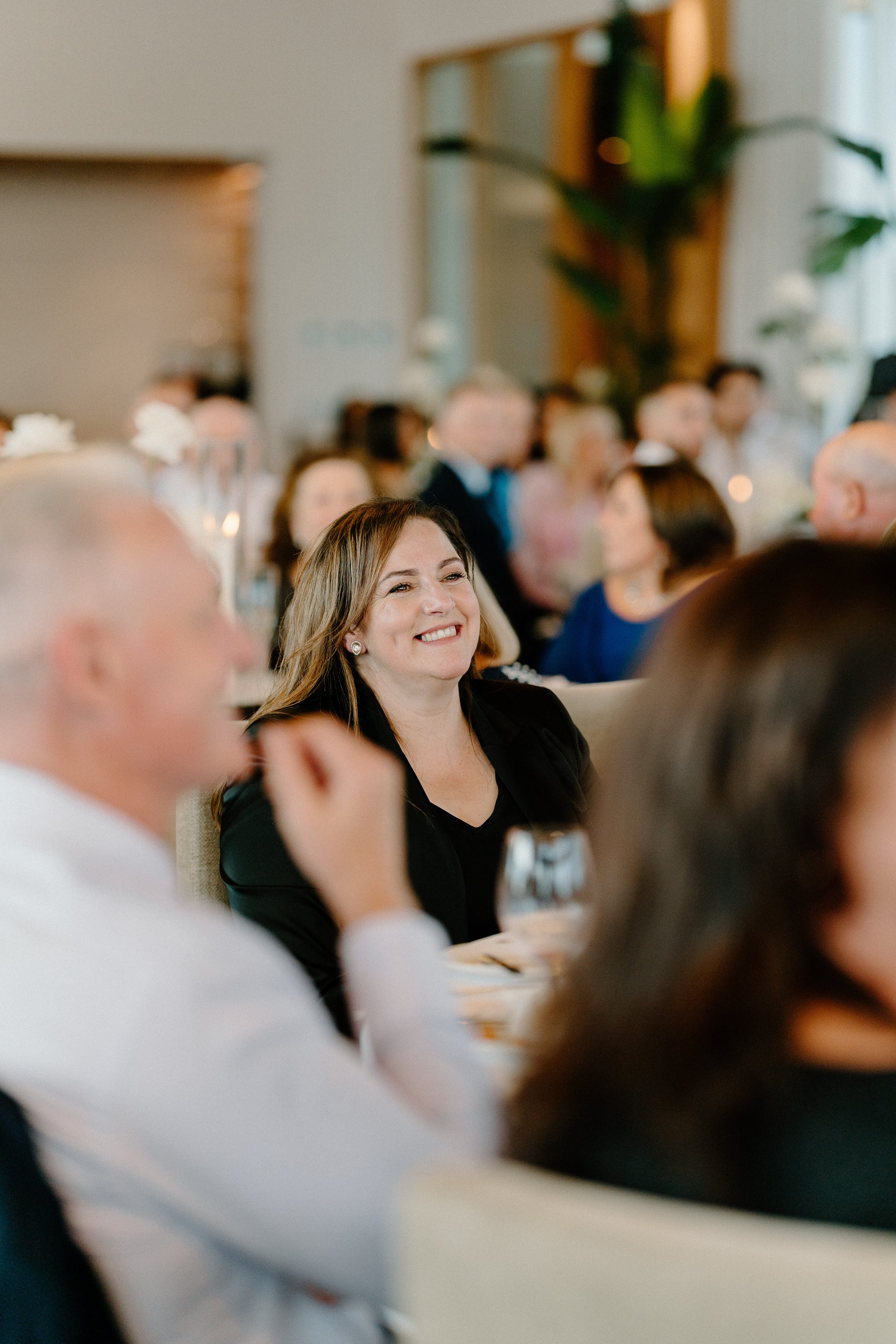 A woman smiling at a formal event with others seated around her, in a well-lit venue with large plants and decorative candles.