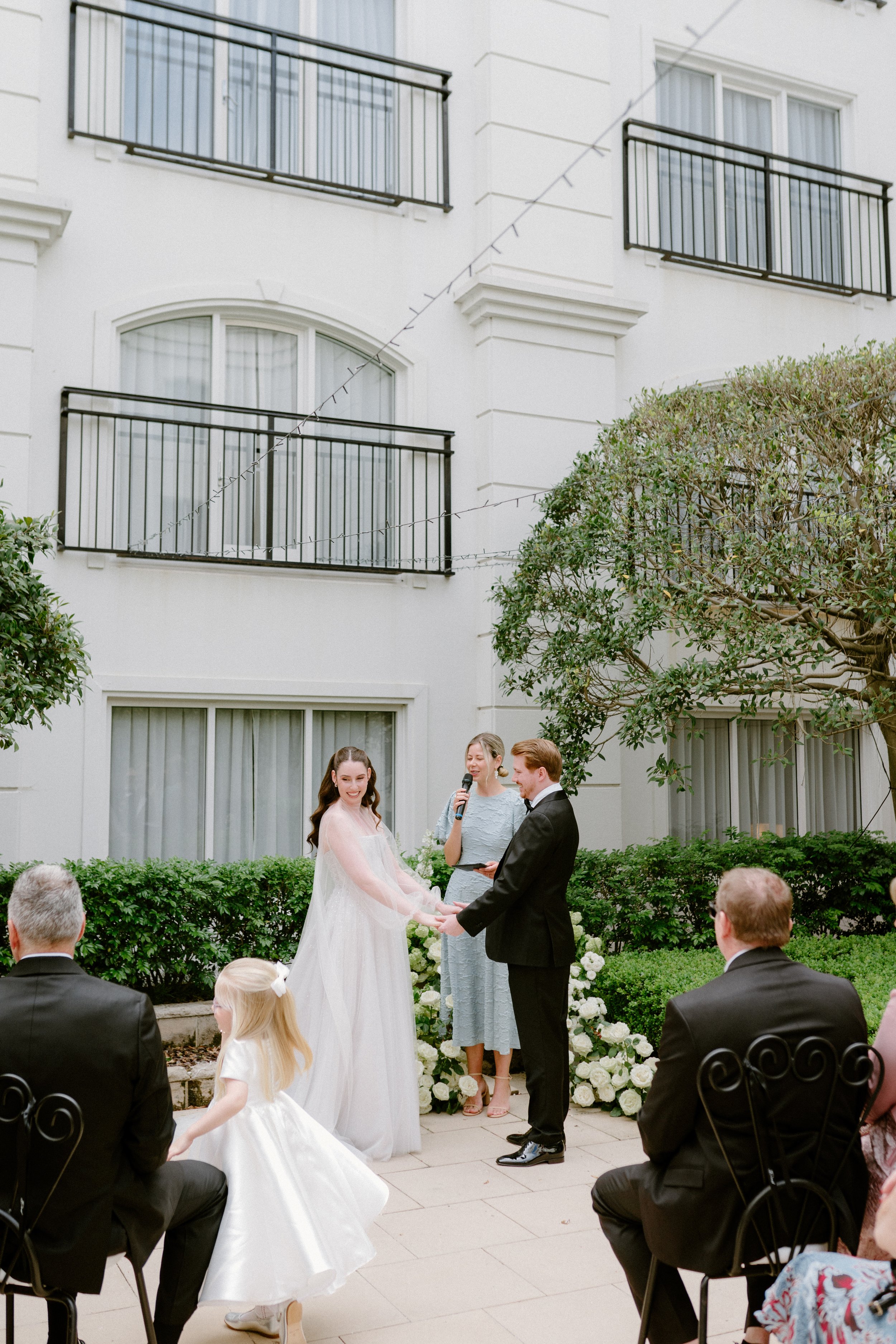 A wedding ceremony outdoors with a bride and groom holding hands, a woman officiating, and guests seated nearby, in an urban garden setting with a white building and greenery.
