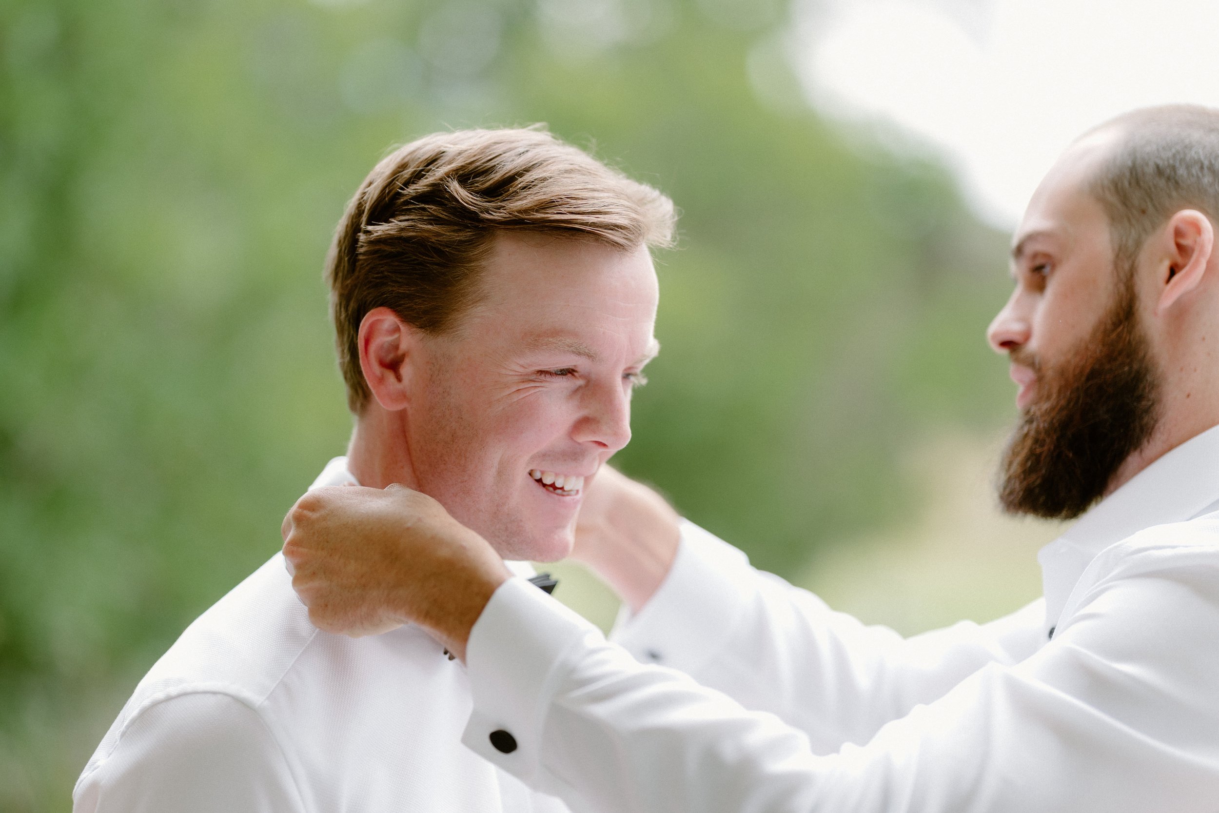 Two men, dressed in white tuxedos, are smiling and laughing while embracing each other outdoors with a blurred green background.