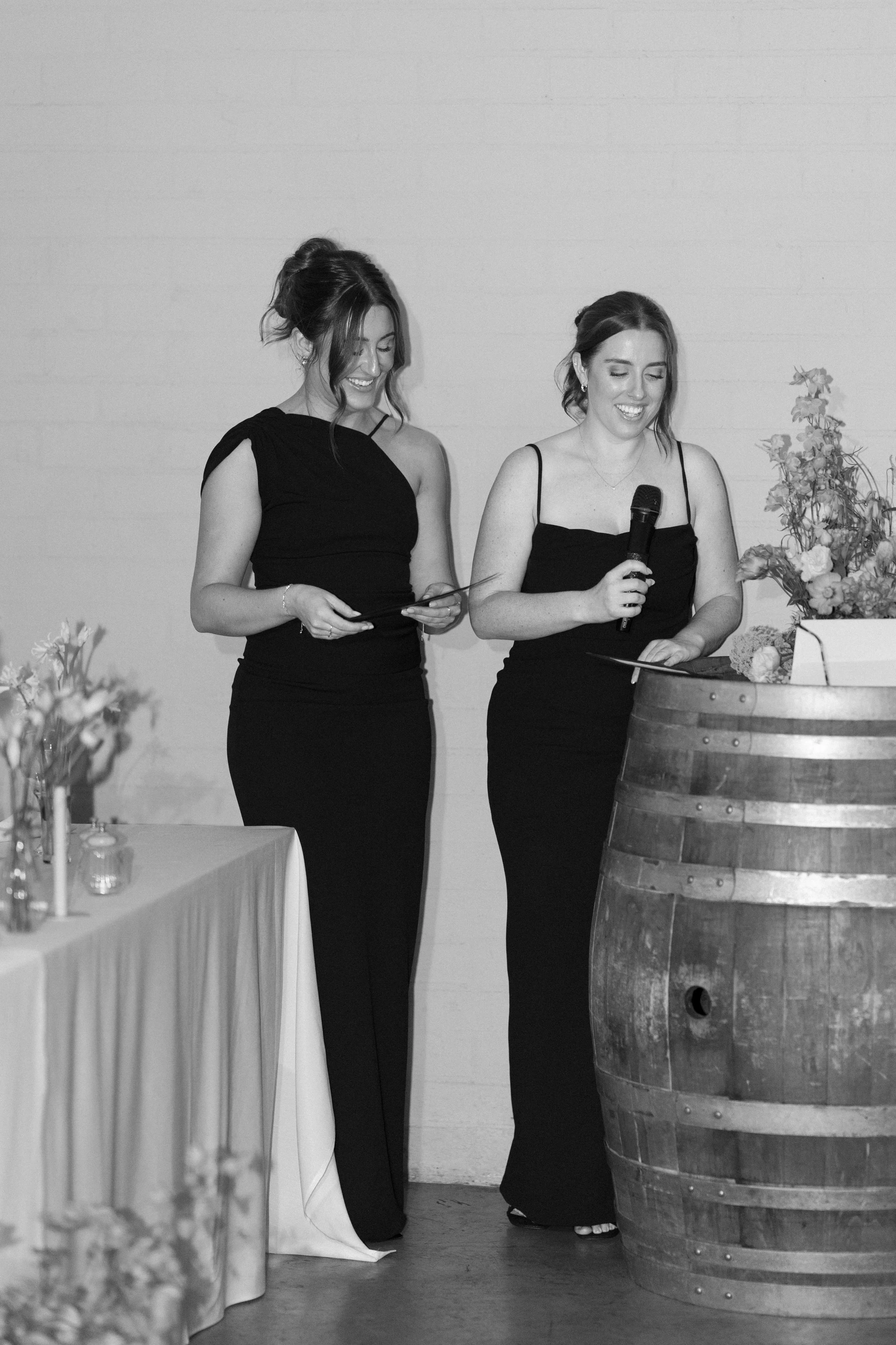 Two women in black dresses smiling, one holding a microphone, standing near a wooden wine barrel with flowers on top, at a celebration or event.