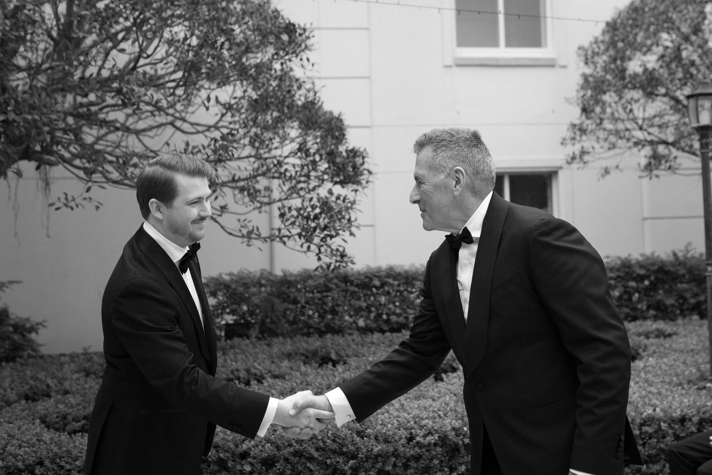 Two men in tuxedos shaking hands outdoors, smiling, with trees and a building in the background.