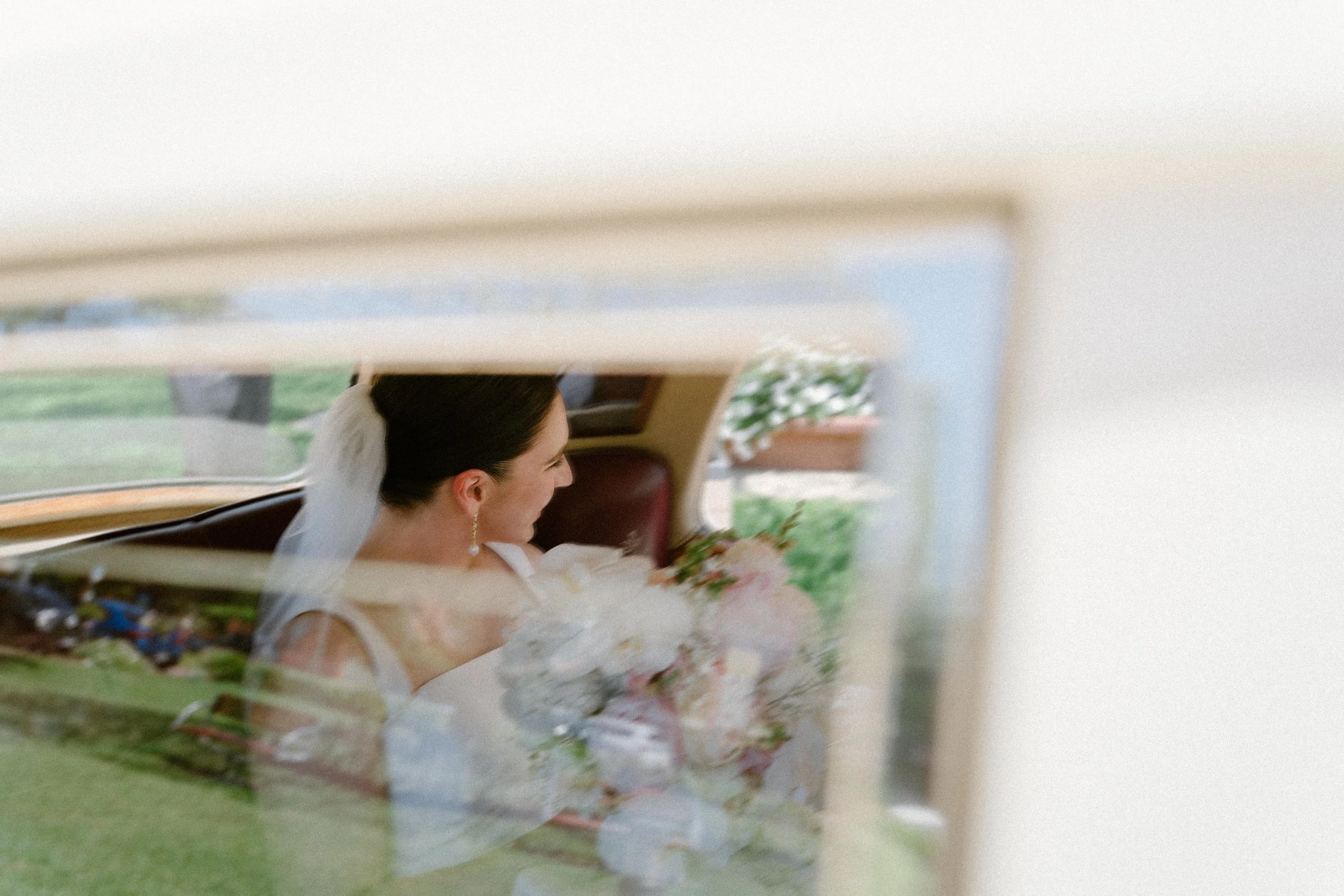 A bride with dark hair and a white veil sitting inside a vintage car, holding a bouquet of flowers, reflected in a window.