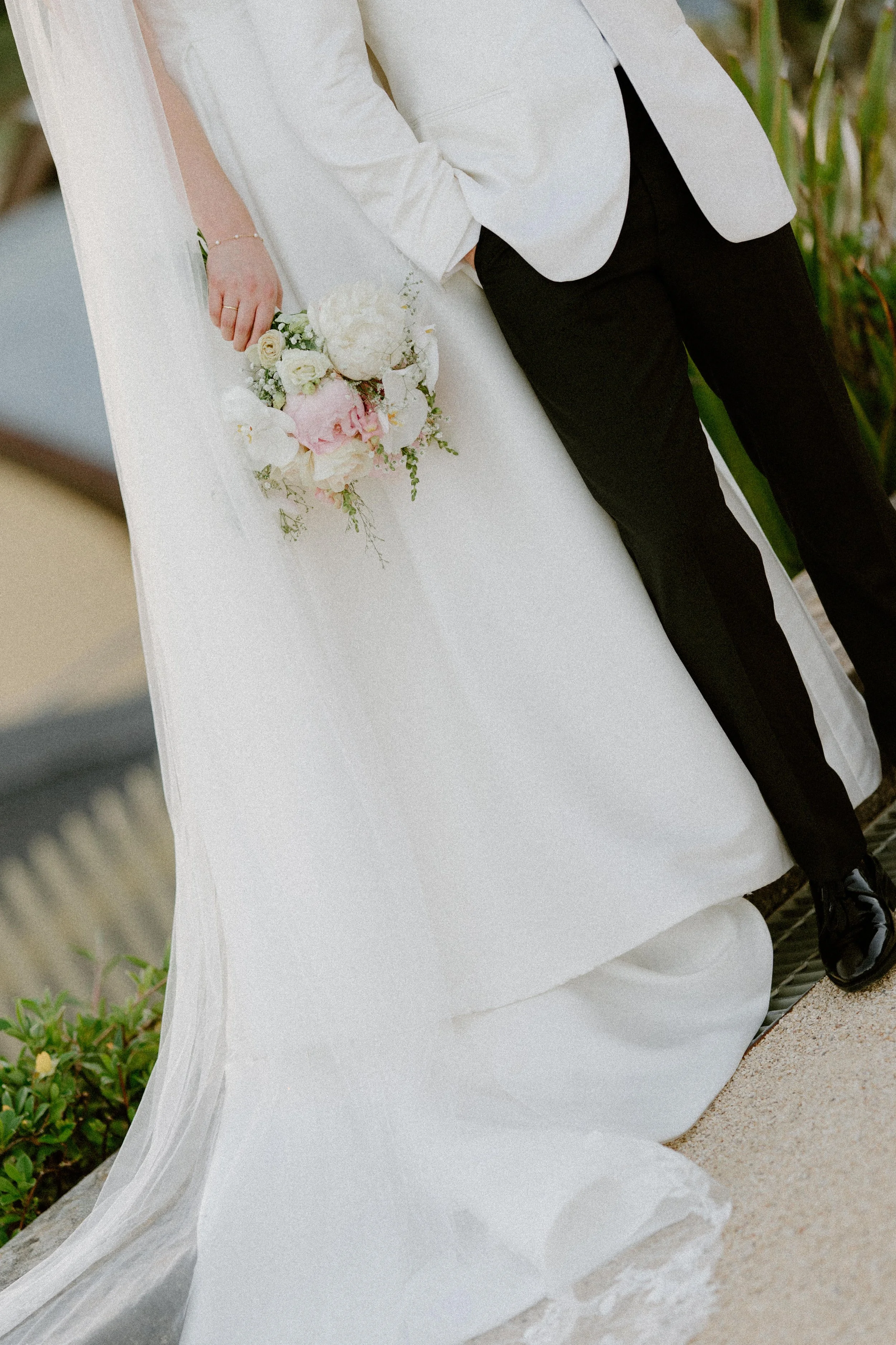 Close-up of a bride and groom at a wedding, with the bride holding a bouquet of white and pink flowers and the groom wearing a white jacket and black pants.