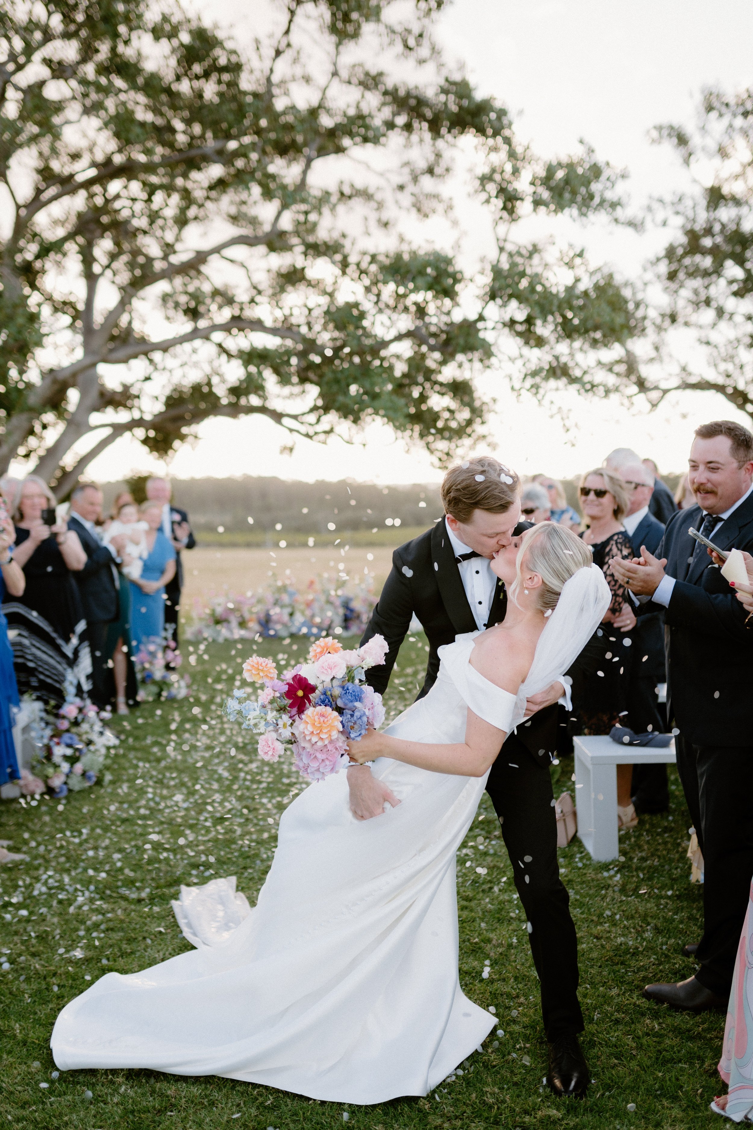 A bride and groom kiss during their outdoor wedding ceremony, surrounded by guests and colorful flowers, with a large tree and open sky in the background. Bimbadgen Palmers Lane Wedding, Hunter Valley