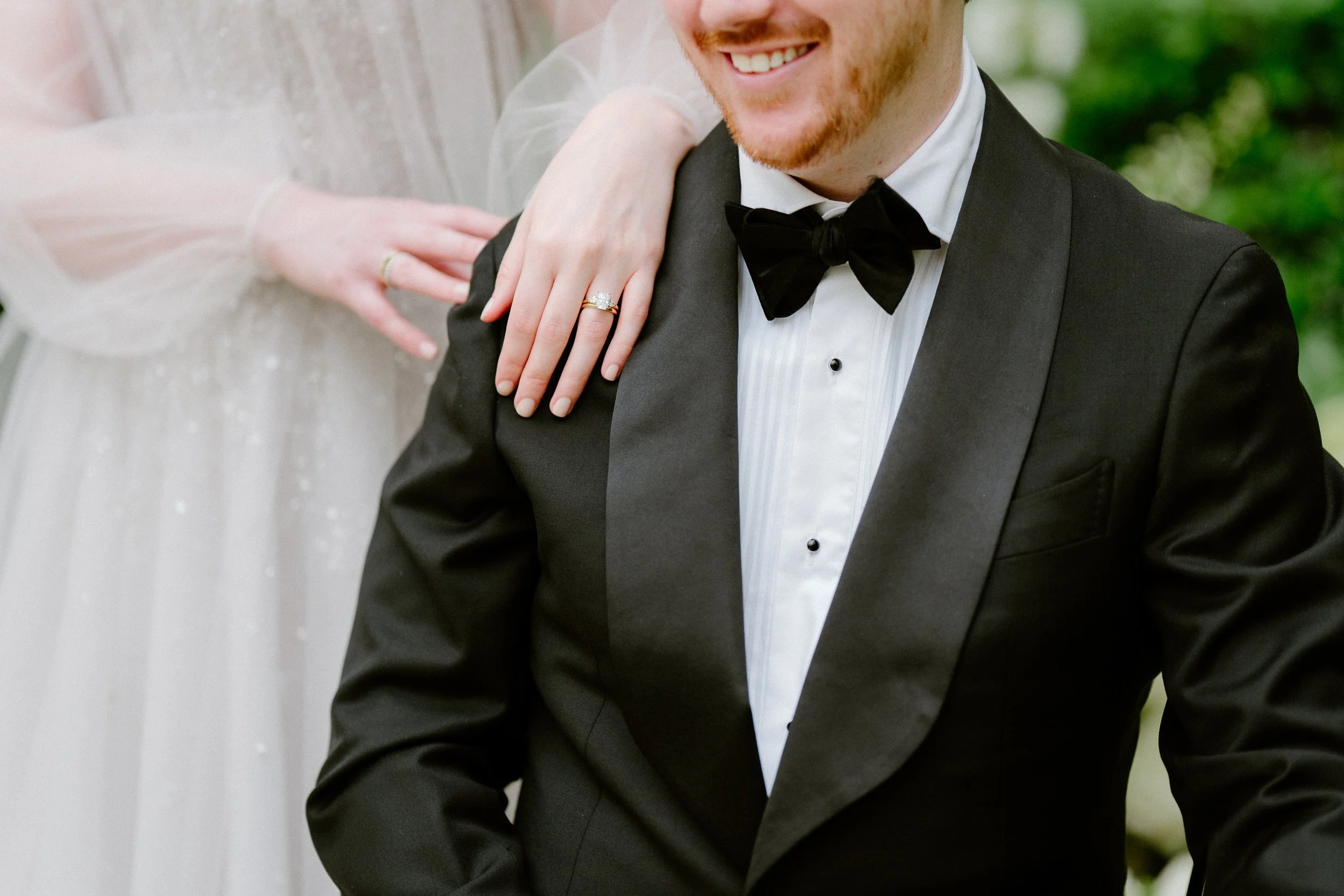 Close-up of a man in a black tuxedo with a bow tie, smiling, with a woman's hand resting on his shoulder. The woman is wearing a wedding dress and a ring, indicating a wedding celebration.