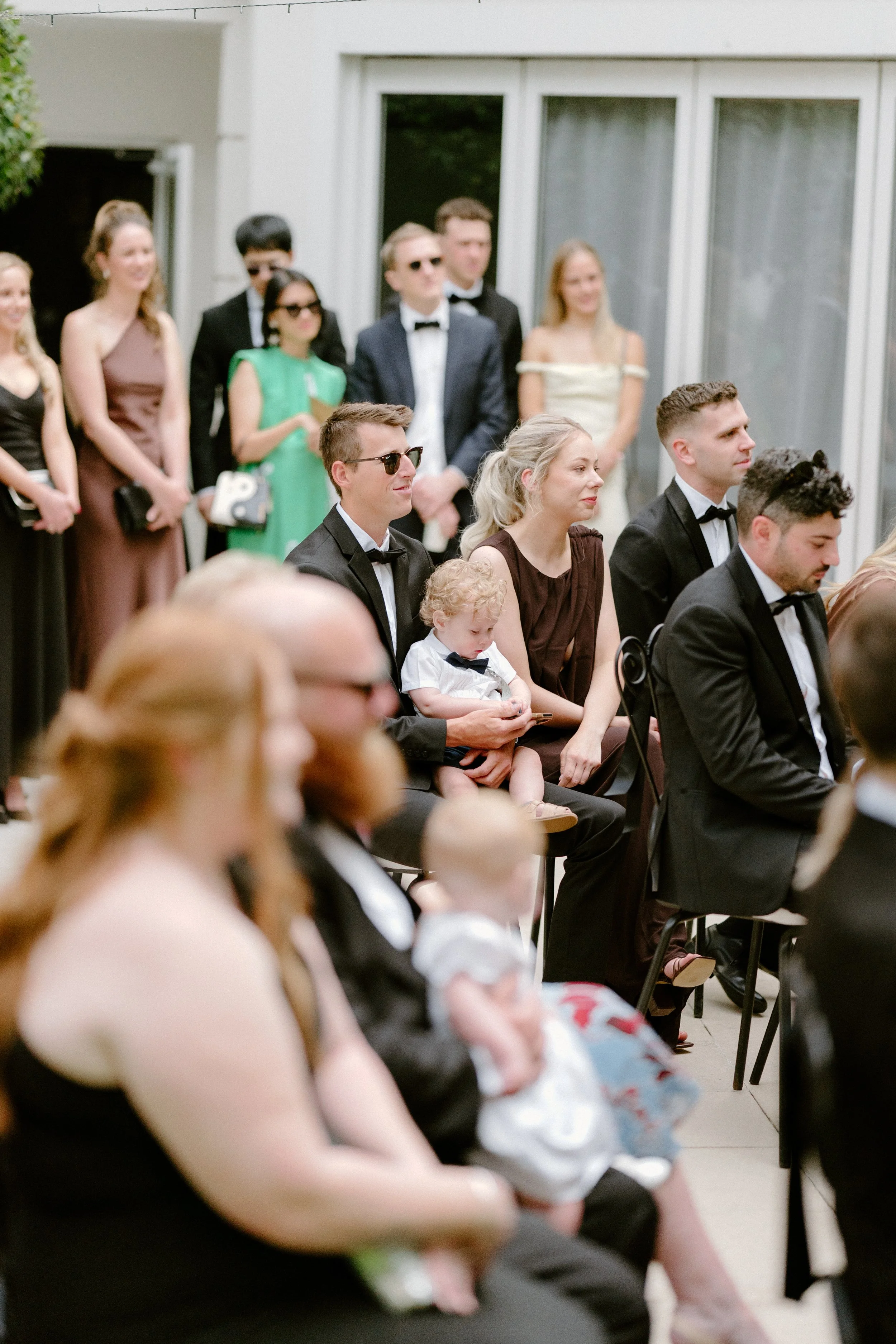 Group of guests attending a formal event, seated and standing, dressed in suits, dresses, and tuxedos, with some wearing sunglasses, indoors near a window.
