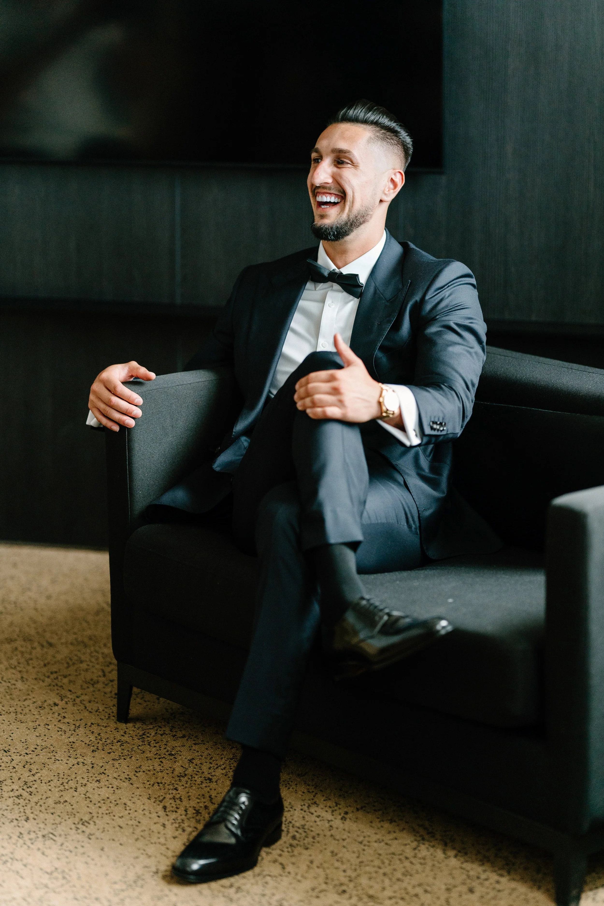 A man in a tuxedo sitting on a black sofa, smiling and giving a thumbs up.