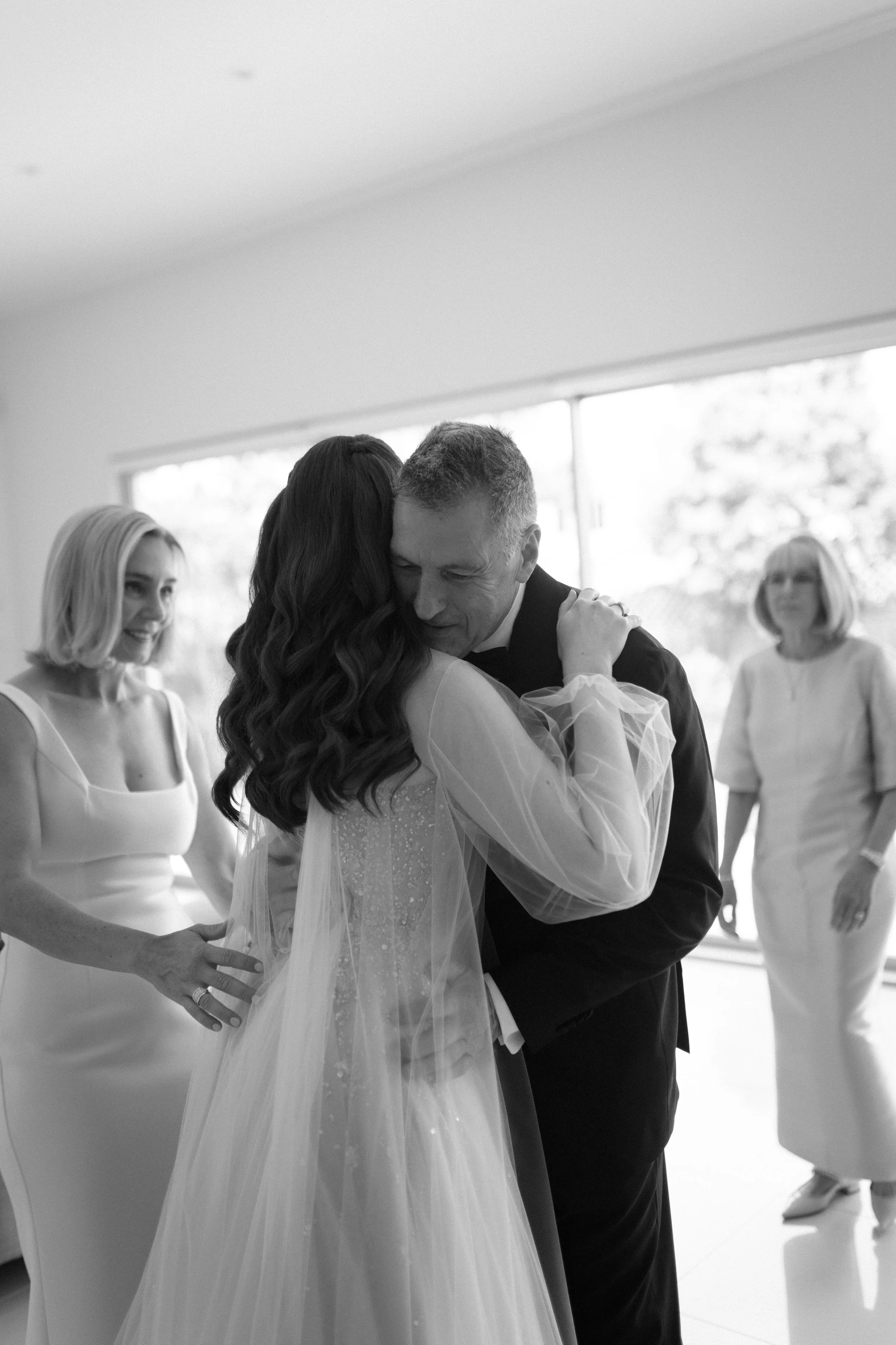 A black and white photo of a wedding dance featuring a bride and groom embracing, with two women watching happily in the background.