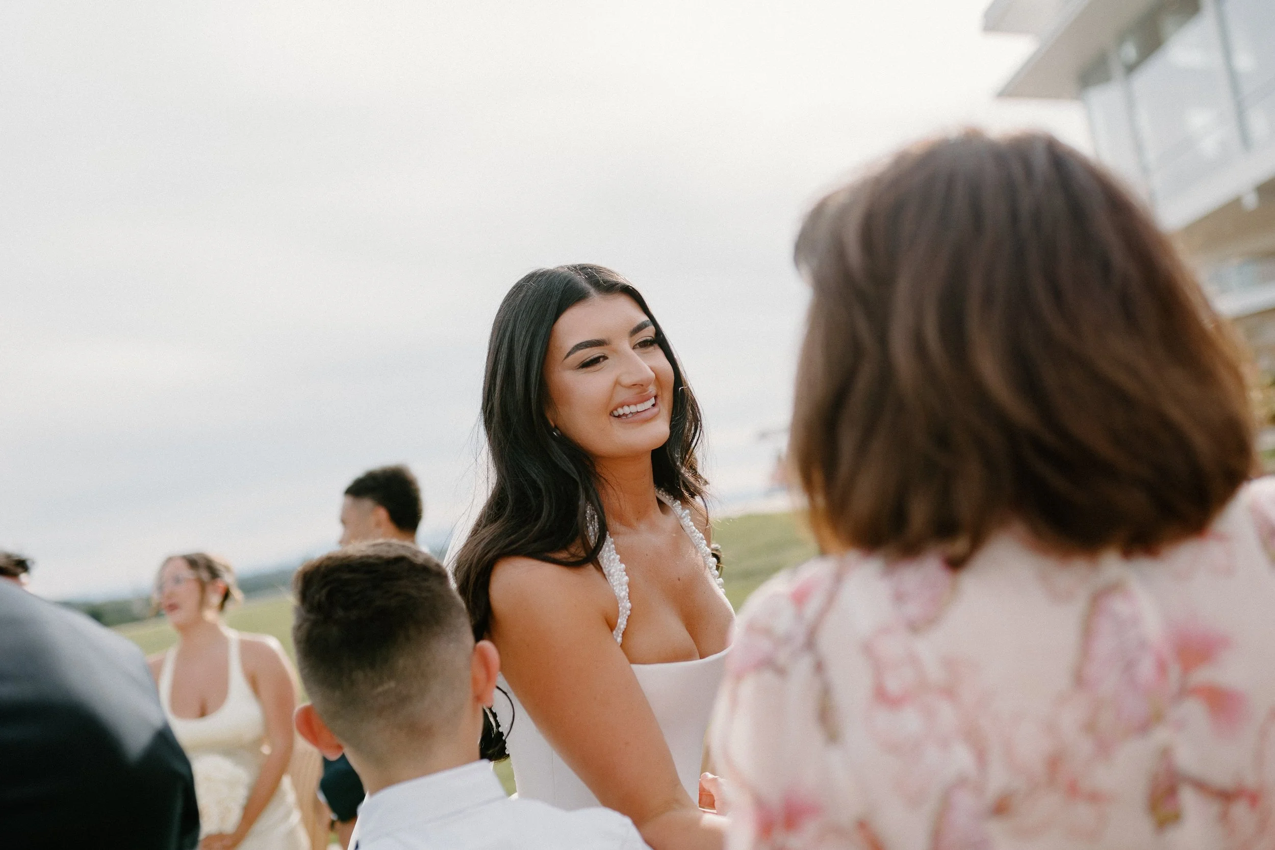 A woman with long black hair smiling at another woman with brown hair during an outdoor event, with several other people in the background and modern buildings nearby.