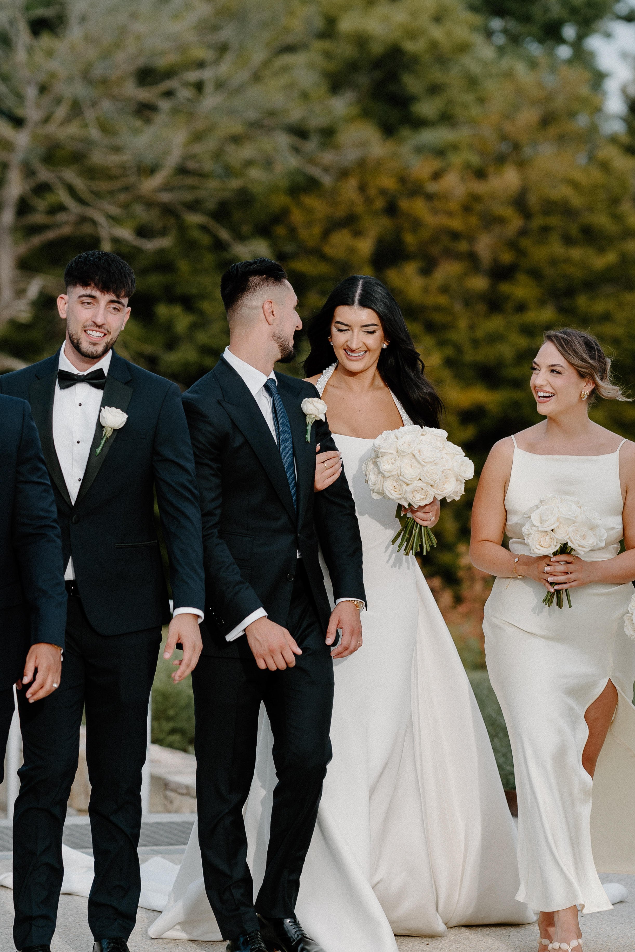 A bride in a white gown holding a bouquet of white roses walking with a groom and friends at a wedding ceremony outdoors with trees in the background.