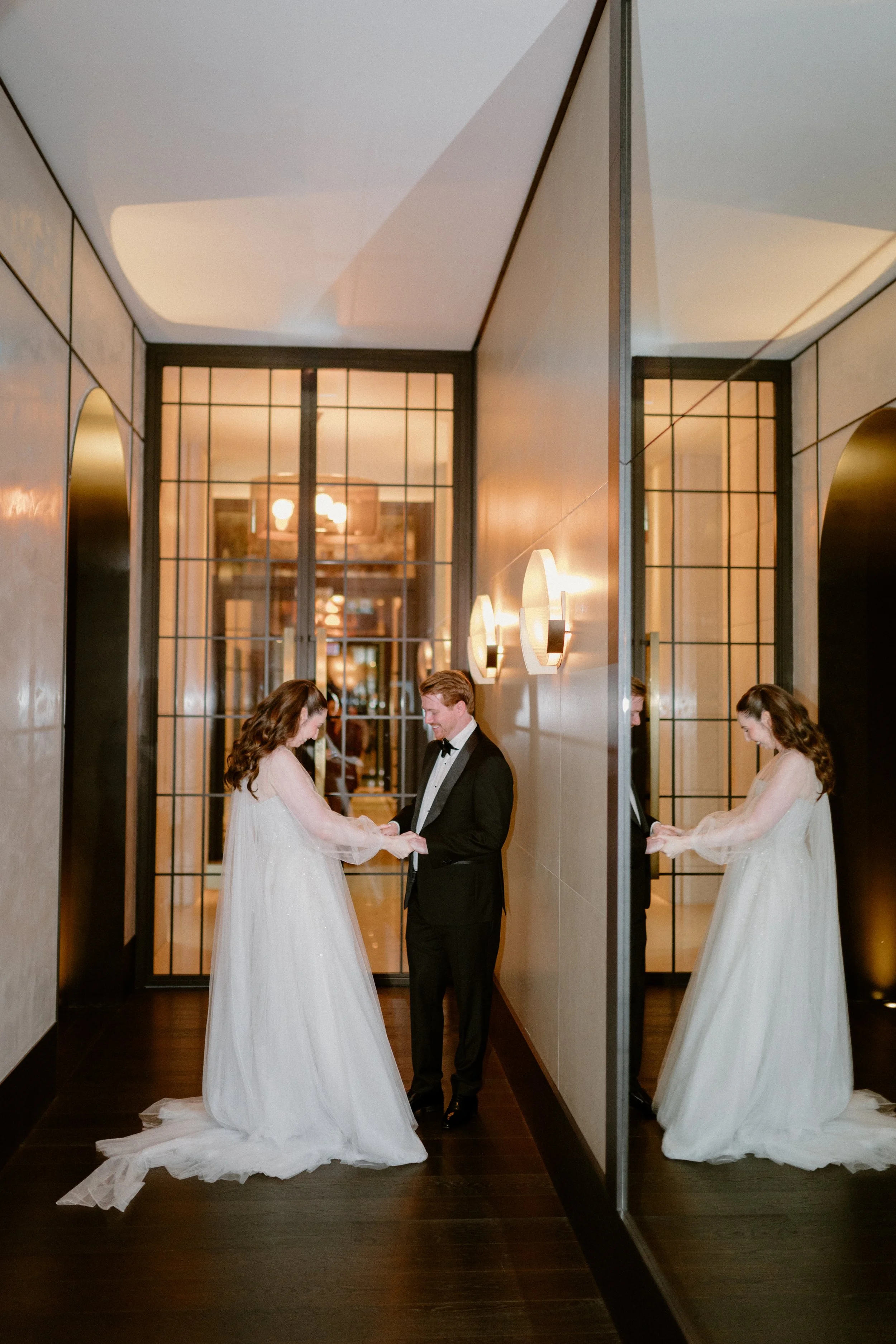 A bride and groom holding hands and smiling at each other in an elegant hallway with large mirrors and warm lighting.