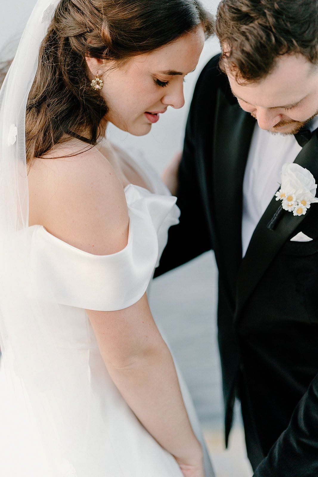 A bride and groom close together, with heads bowed, during their wedding ceremony. The bride is wearing a white off-shoulder wedding dress with a veil and pearl earrings. The groom is dressed in a black tuxedo with a white shirt and a boutonniere of 