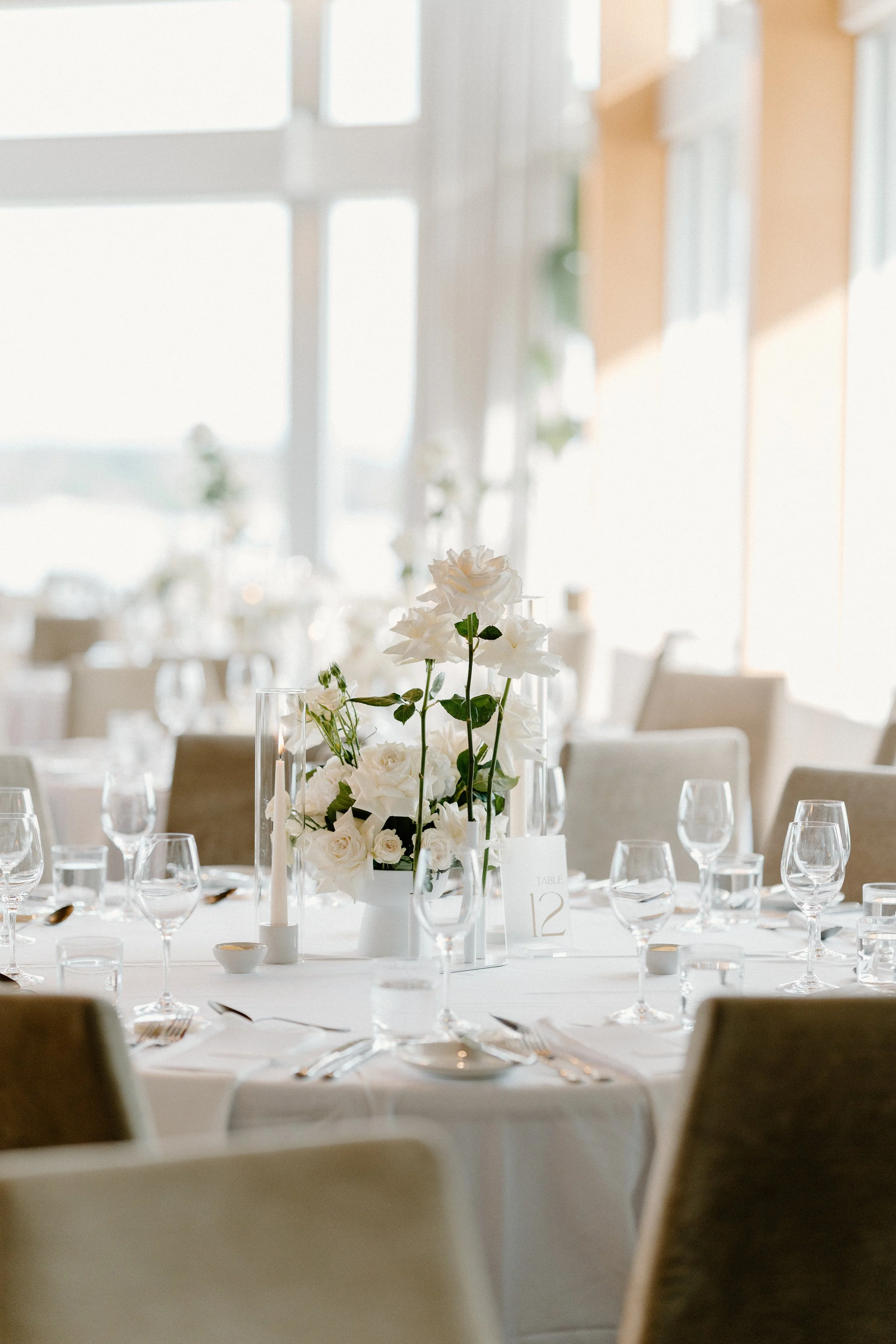 Table setup with white floral centerpiece, surrounded by wine glasses, silverware, and a table number card that reads 'Table 12', in a bright, elegant room.