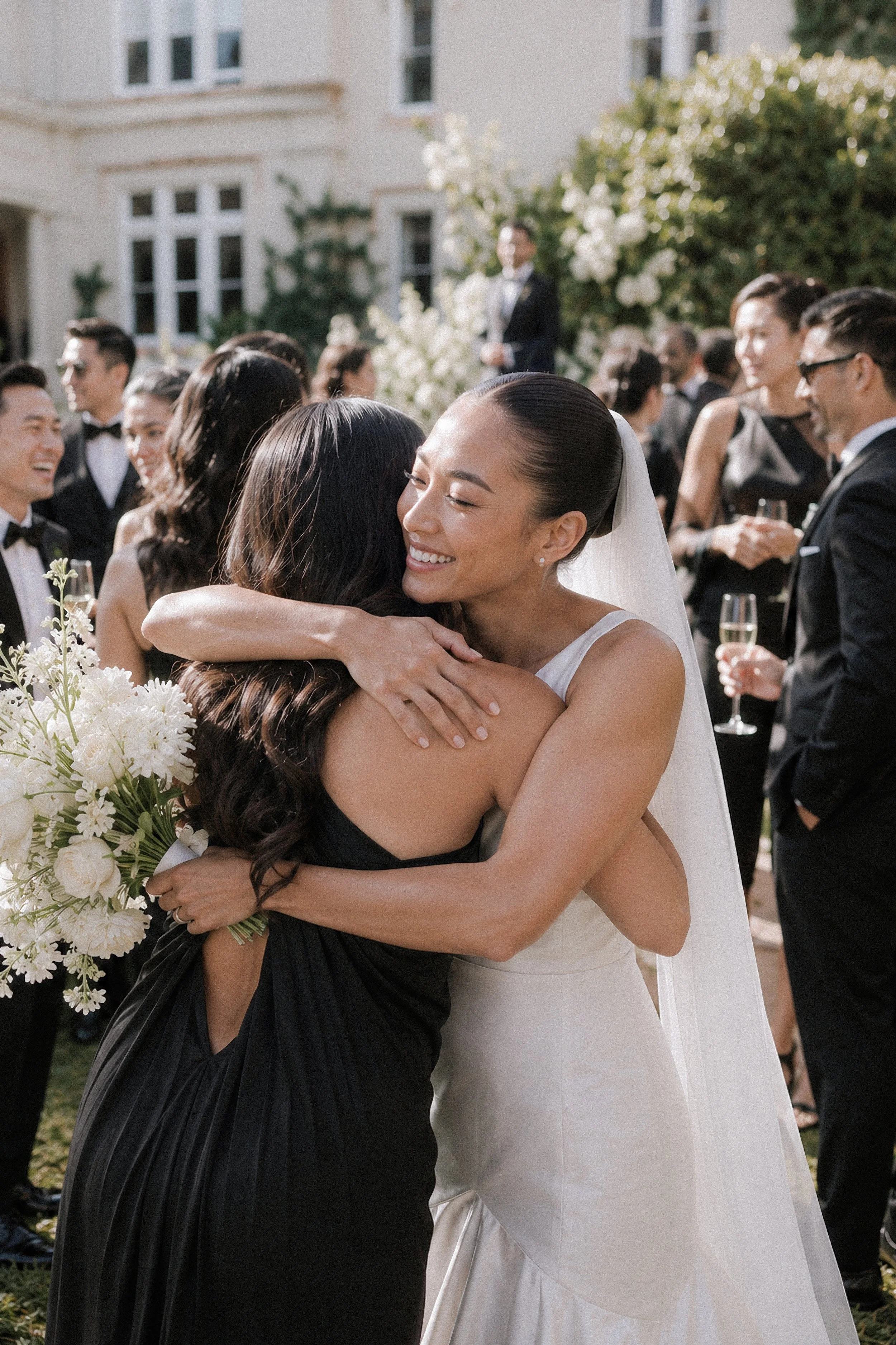 A bride in a white wedding dress hugging a woman in a black dress at an outdoor wedding reception, with guests in formal attire in the background.