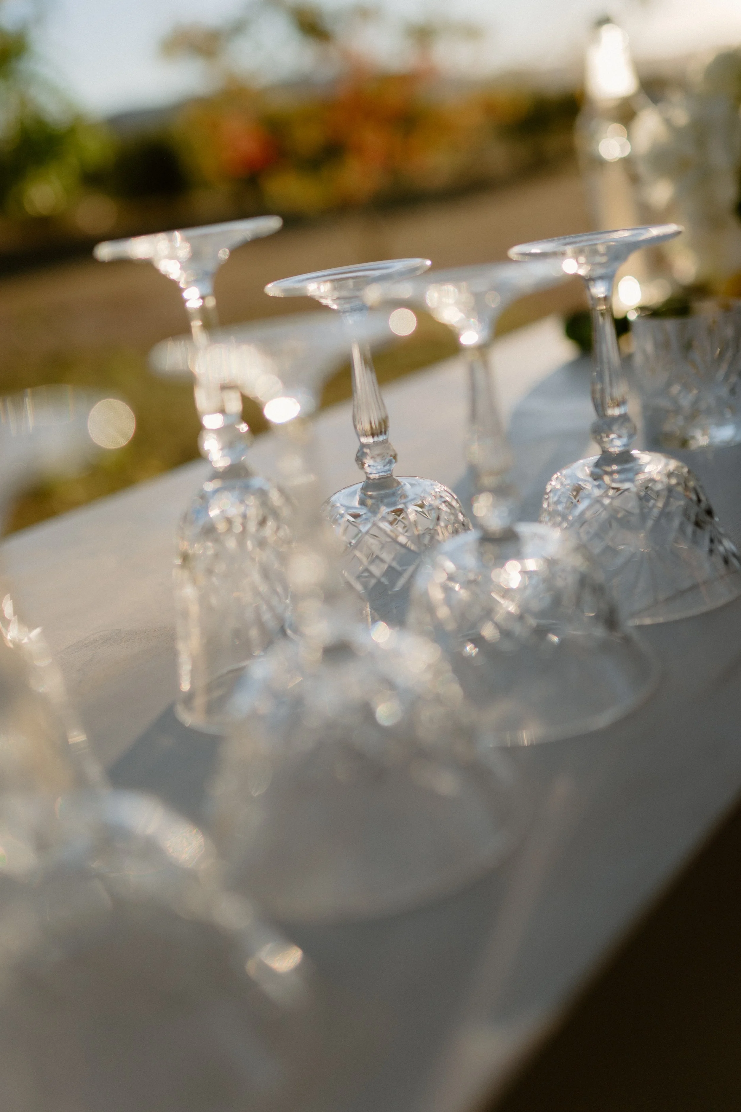 Several upside down glass wine glasses with decorative cuts on the stems, placed on a white surface outdoors during daylight, with blurred trees and sunlight in the background.
