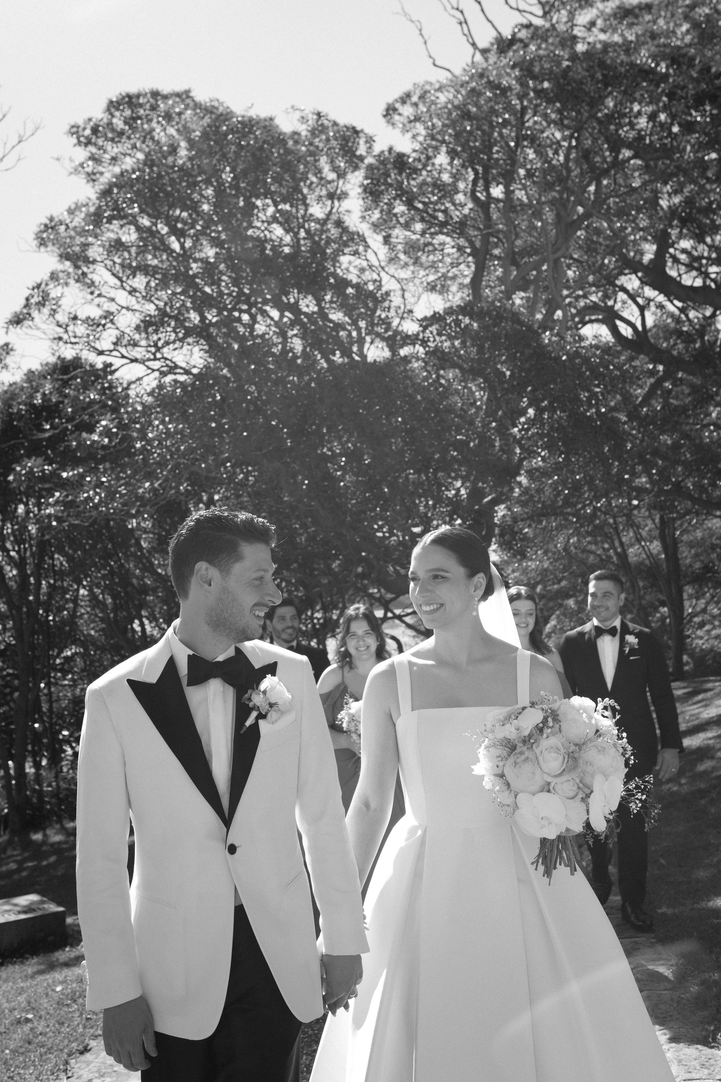 Black and white photo of a newlywed couple walking outdoors, holding hands. The groom is in a tuxedo with a bow tie, and the bride is in a white wedding dress holding a bouquet. They are smiling at each other, with guests in the background, surrounde