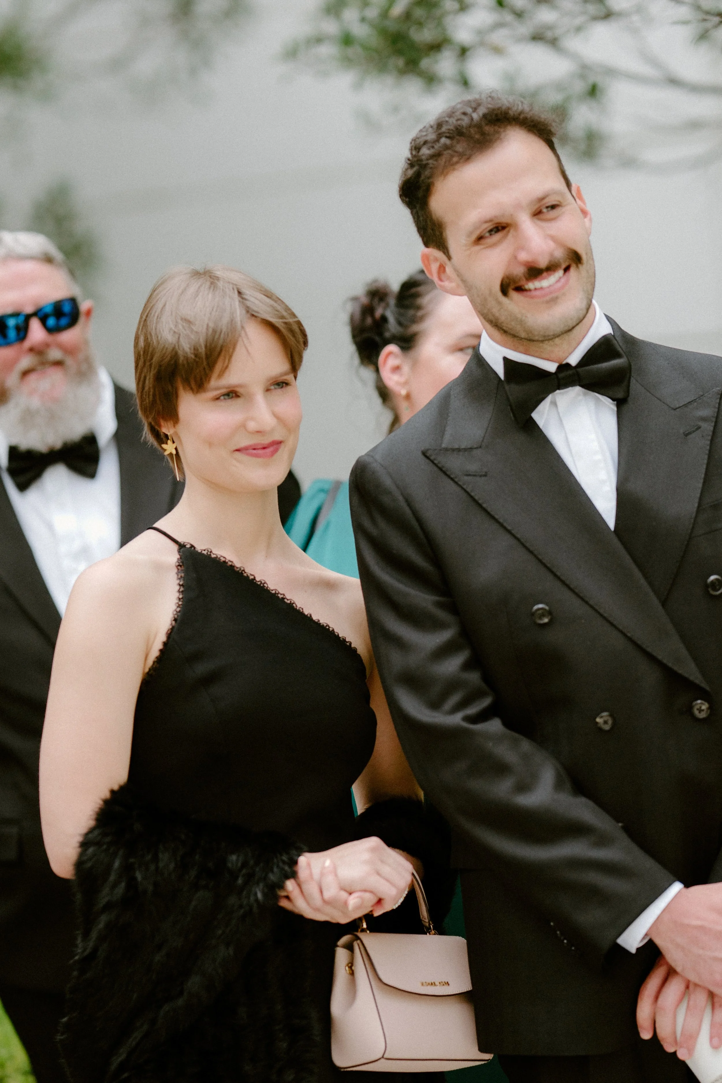 Group of people at a formal outdoor event, with a man in a tuxedo smiling in the foreground and a woman in a black dress standing behind him, holding a beige purse, with other guests in the background near a body of water.