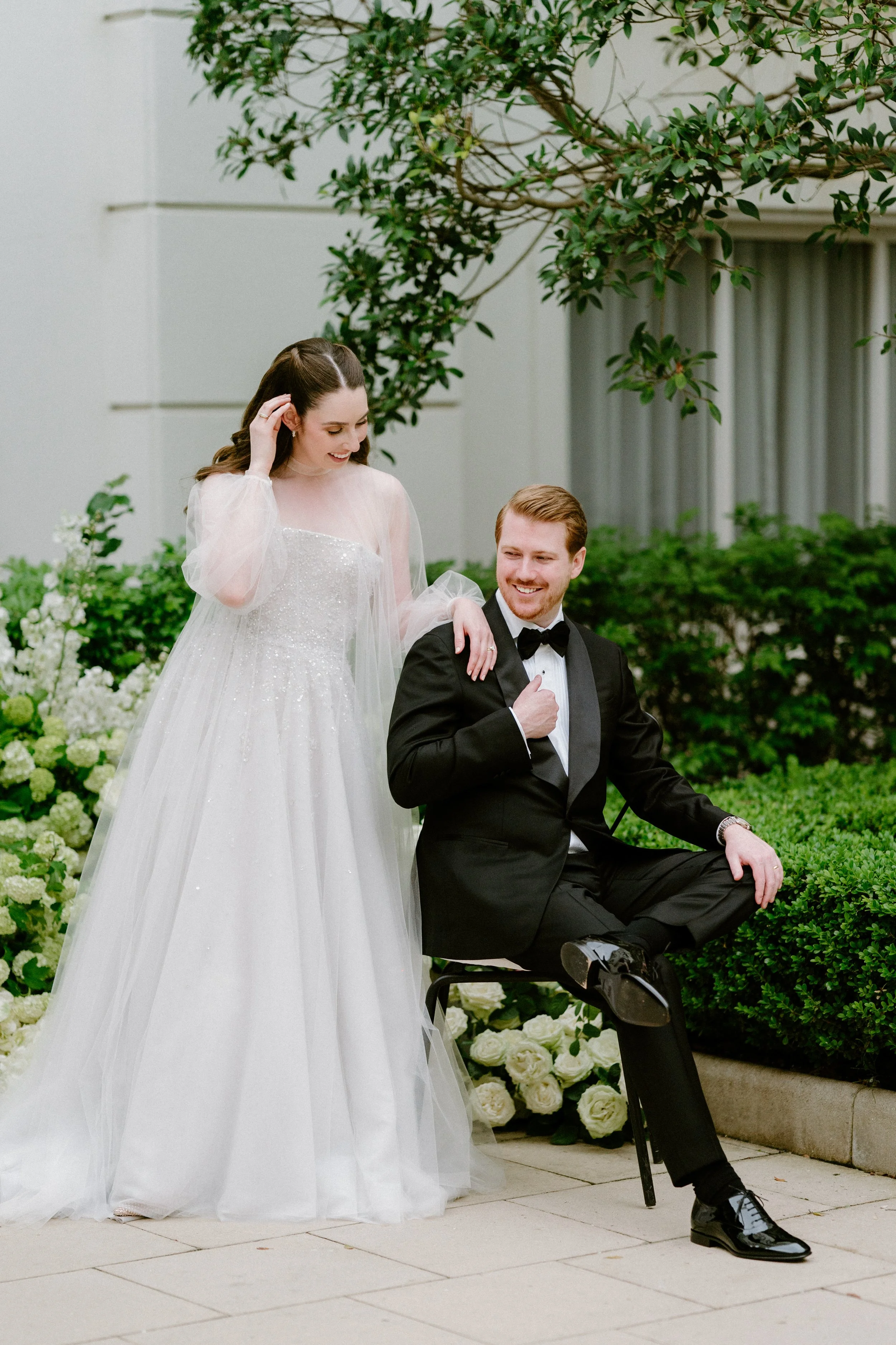 A bride and groom posing together outdoors, with the bride standing and the groom sitting on a chair, surrounded by greenery and white flowers.
