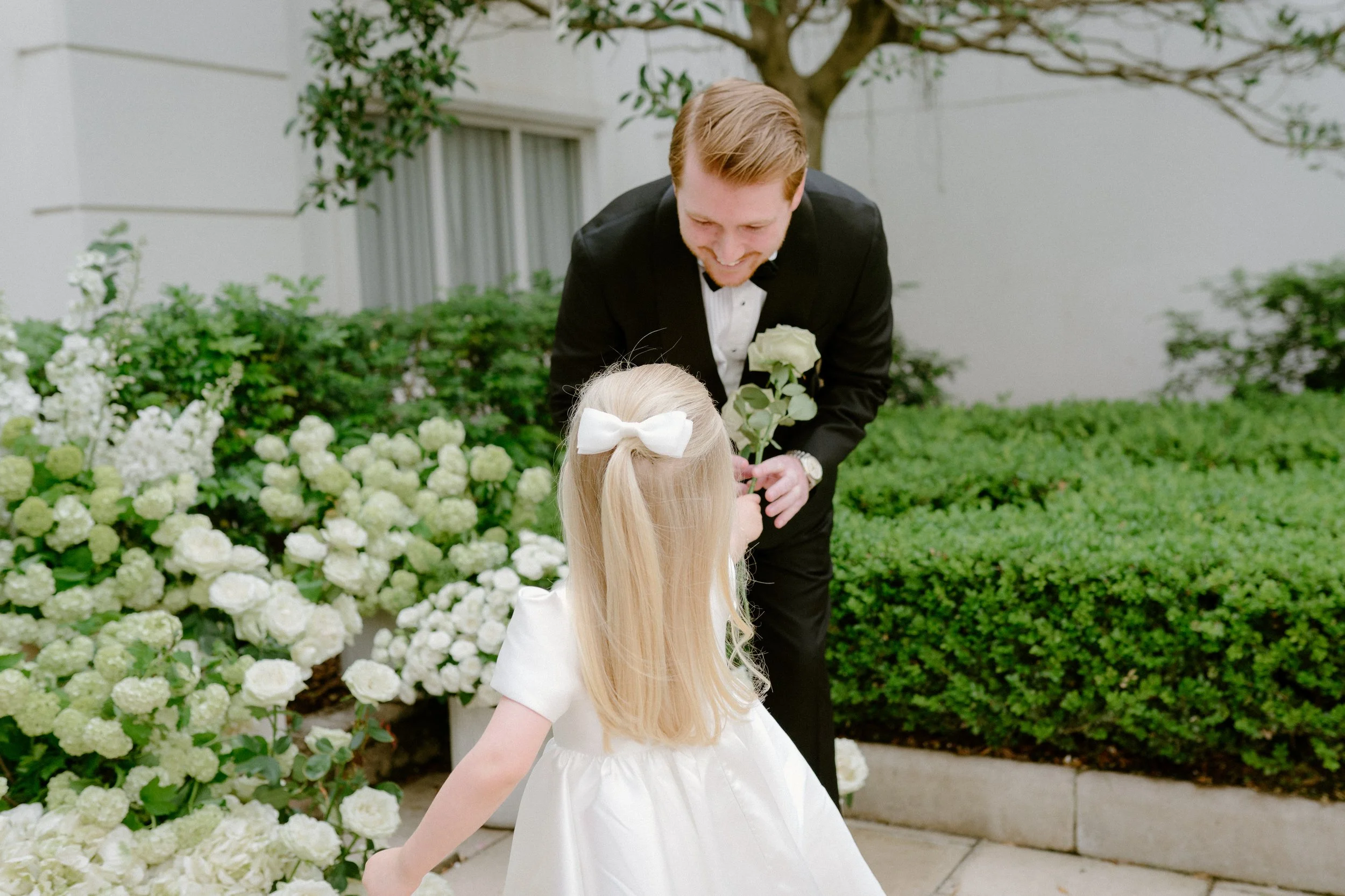 A man in a black tuxedo bending down and smiling at a young girl with long blonde hair, wearing a white dress and a white bow, in a garden with white flowers and green bushes.