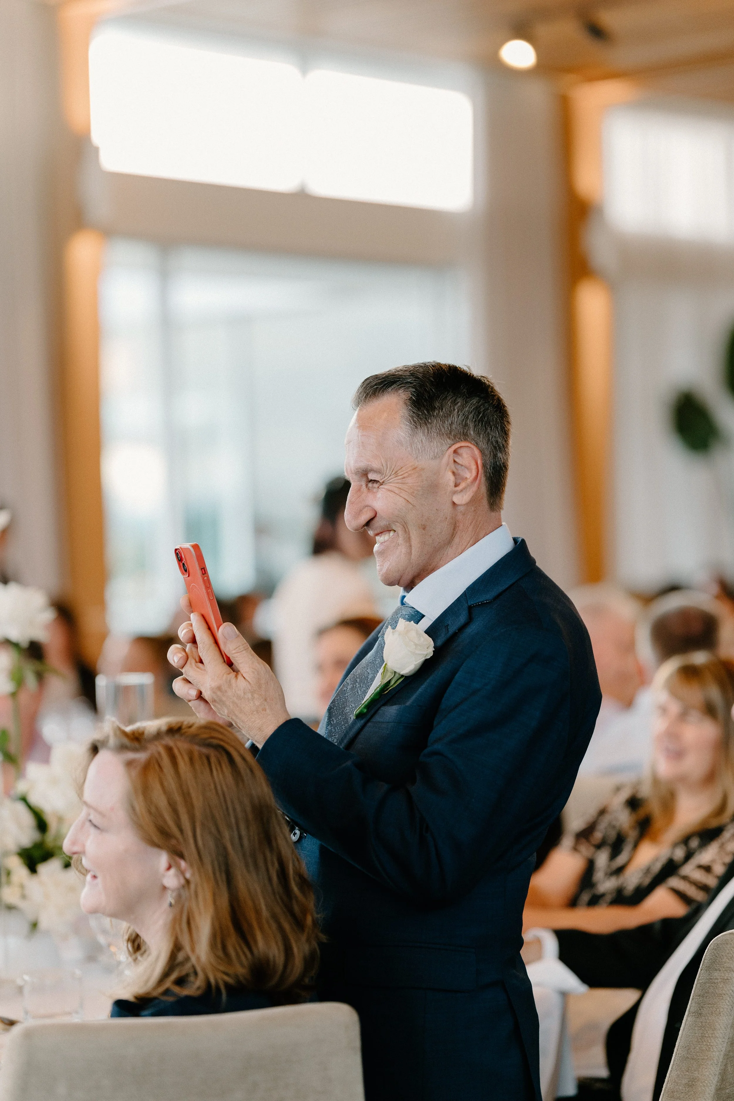 An older man in a dark suit is smiling and taking a photo with his phone at a wedding reception. He has a white flower boutonniere on his suit. Guests are sitting at tables with floral centerpieces, and the setting is a bright, modern indoor space.