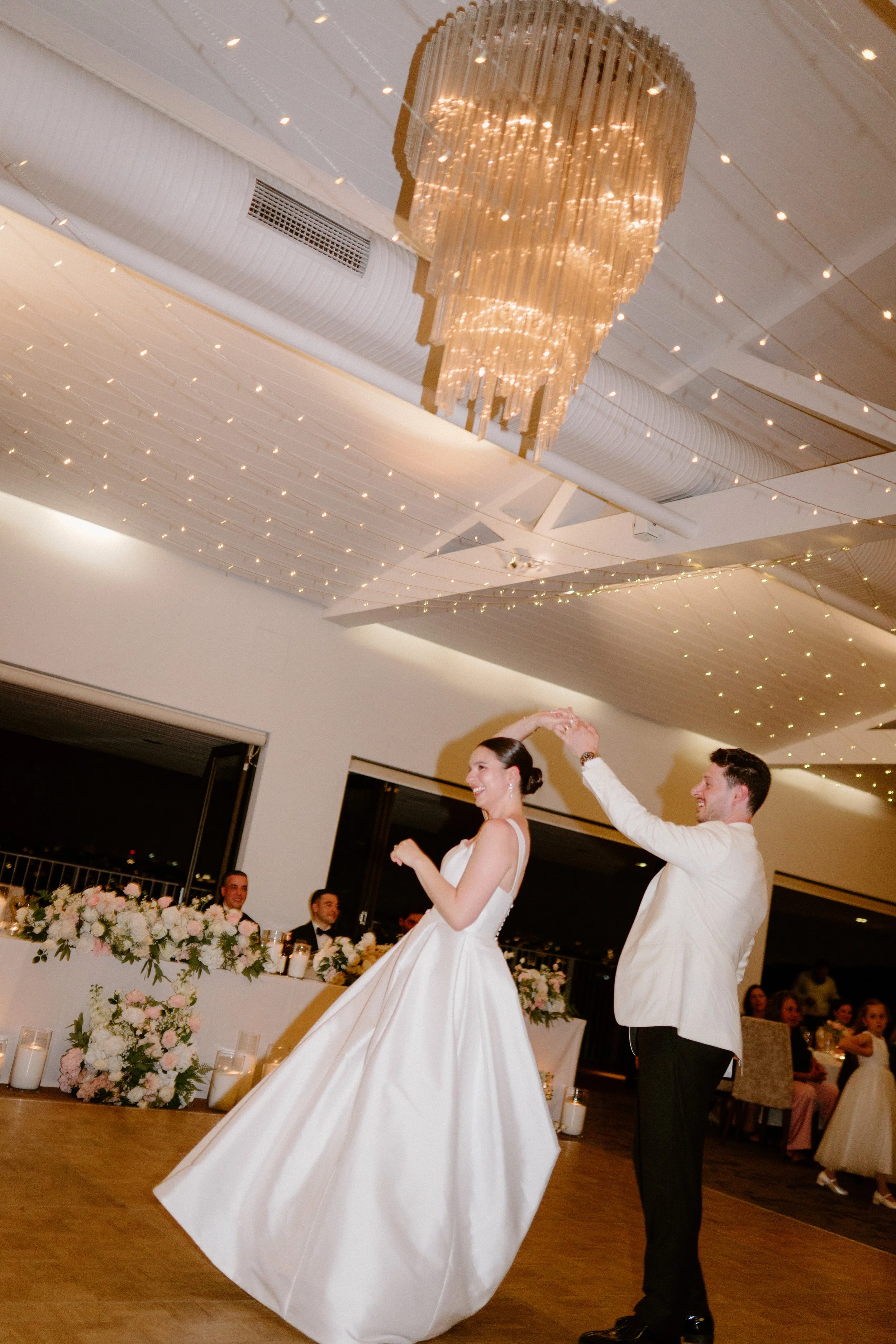 A bride and groom dance at their wedding reception, with the bride twirling in a white gown and the groom in a white jacket and dark pants. The decorated reception hall features floral arrangements and candles, with string lights and an elegant chand