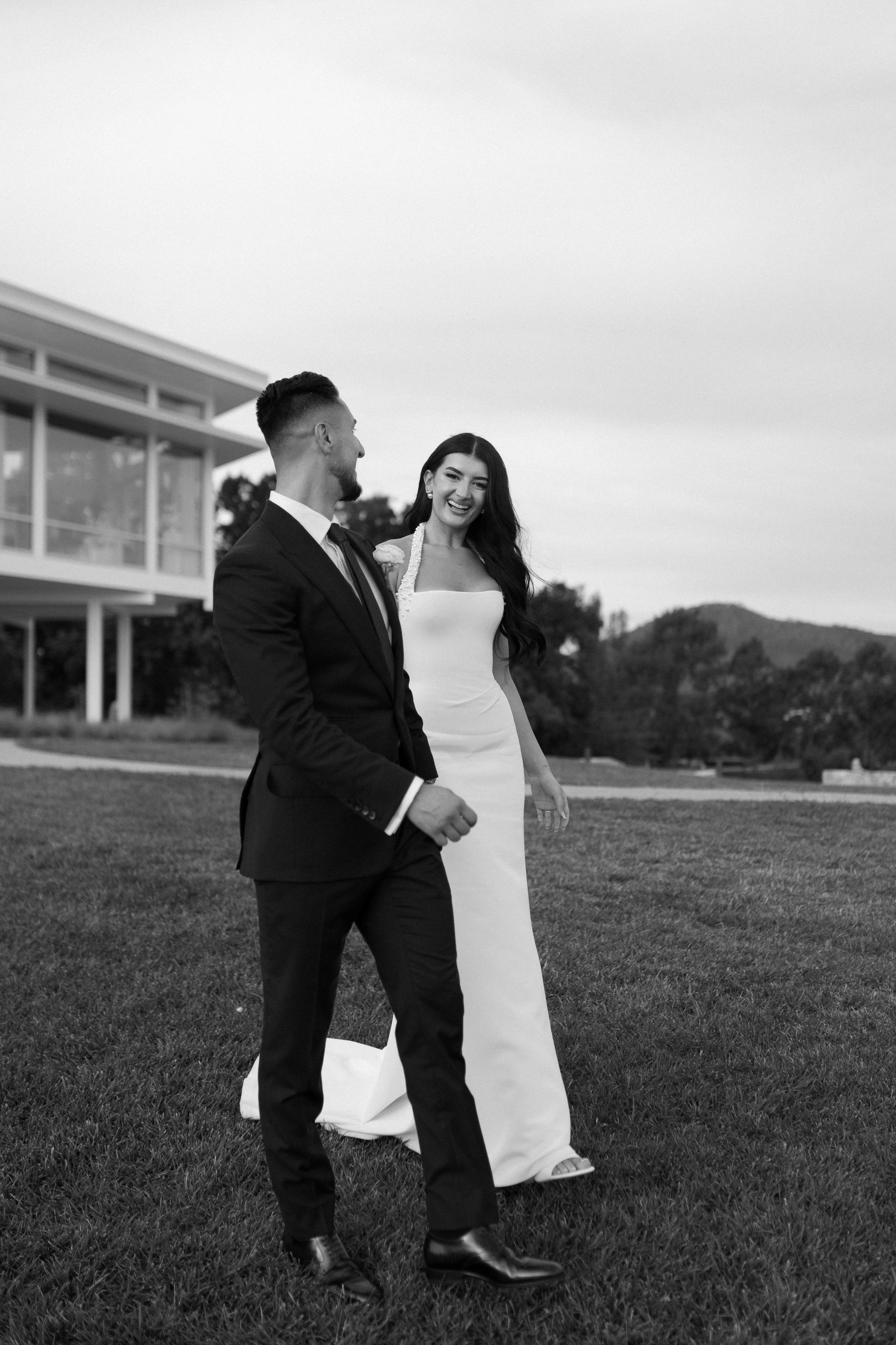 A black and white photo of a smiling woman in a wedding dress walking with a man in a suit on a lawn with modern house and hills in the background.