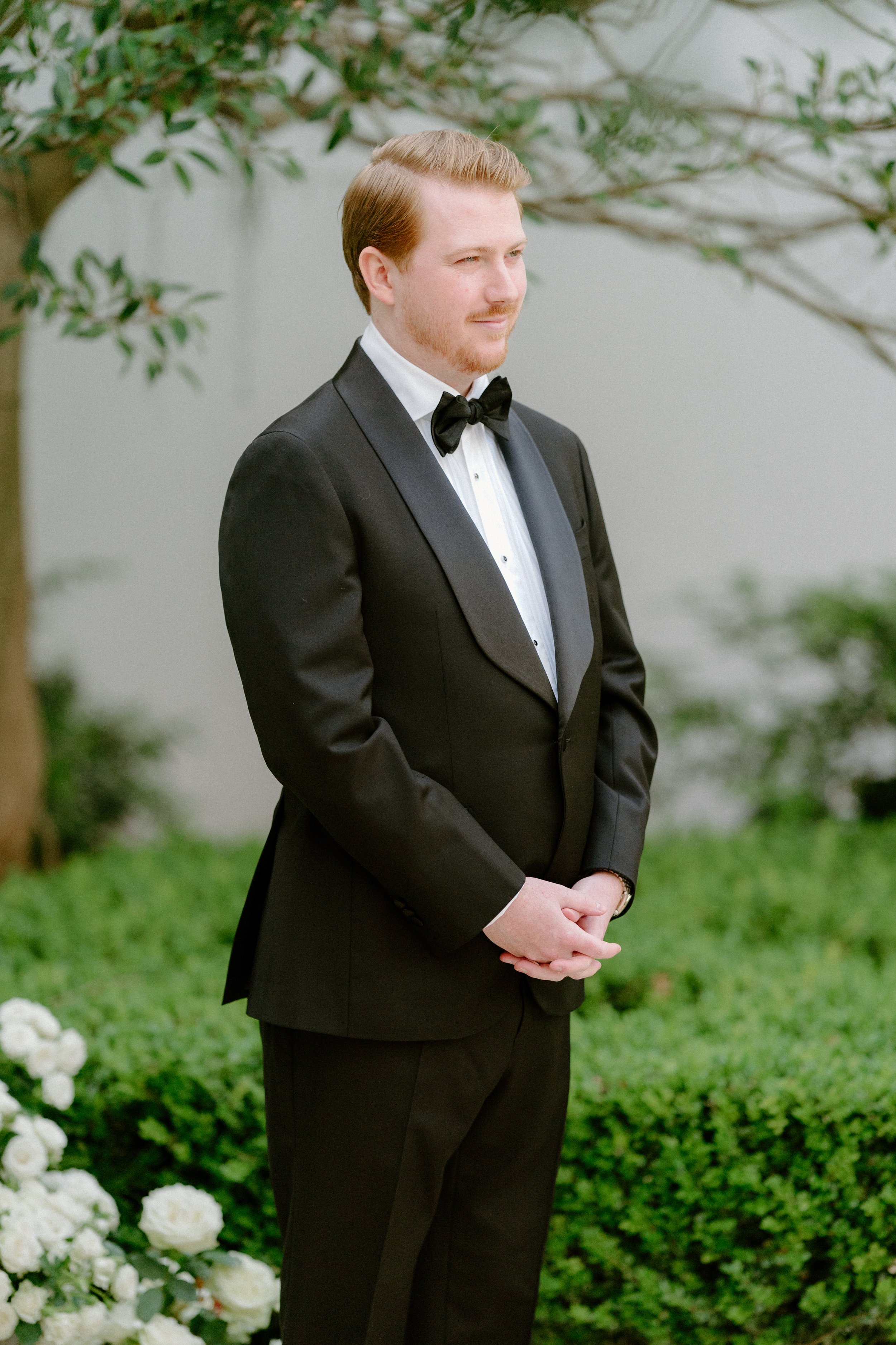 A young man in a black tuxedo with a bow tie stands outdoors near greenery and white flowers, appearing to be at a formal event or wedding.
