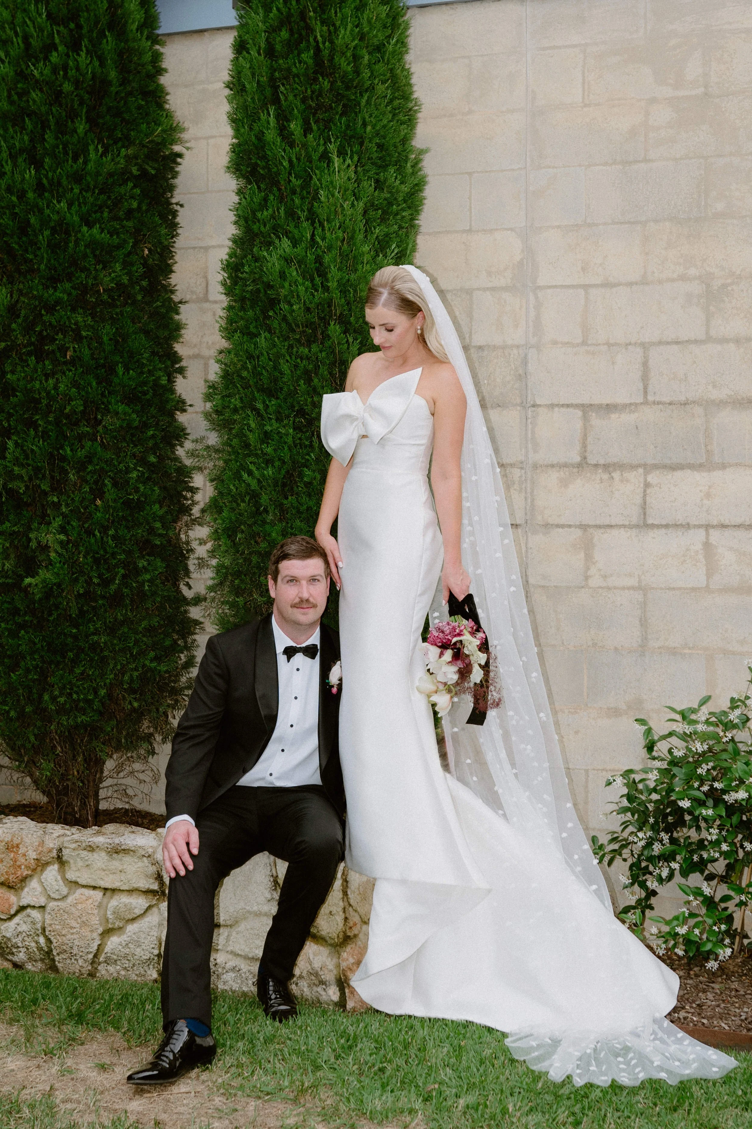 The Sienna, Hunter Valley Wedding Venue, Wedding Photos. A bride in a white wedding dress with a large bow on the bodice and a long veil stands beside a groom in a black tuxedo with a bow tie, both outdoors near green bushes and a stone wall.