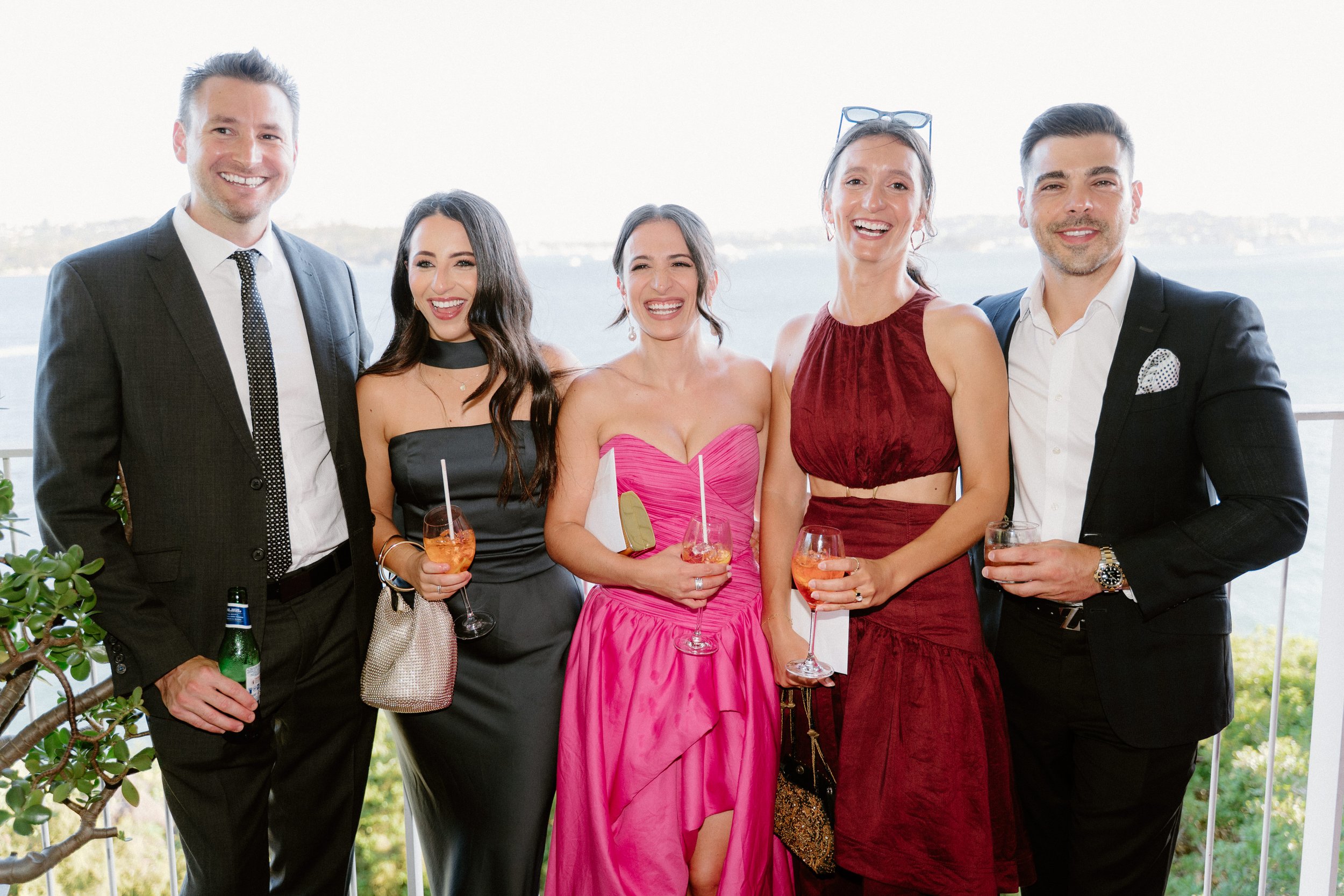 Group of six people in formal attire smiling and holding drinks at an outdoor event with a view of water and trees in the background.