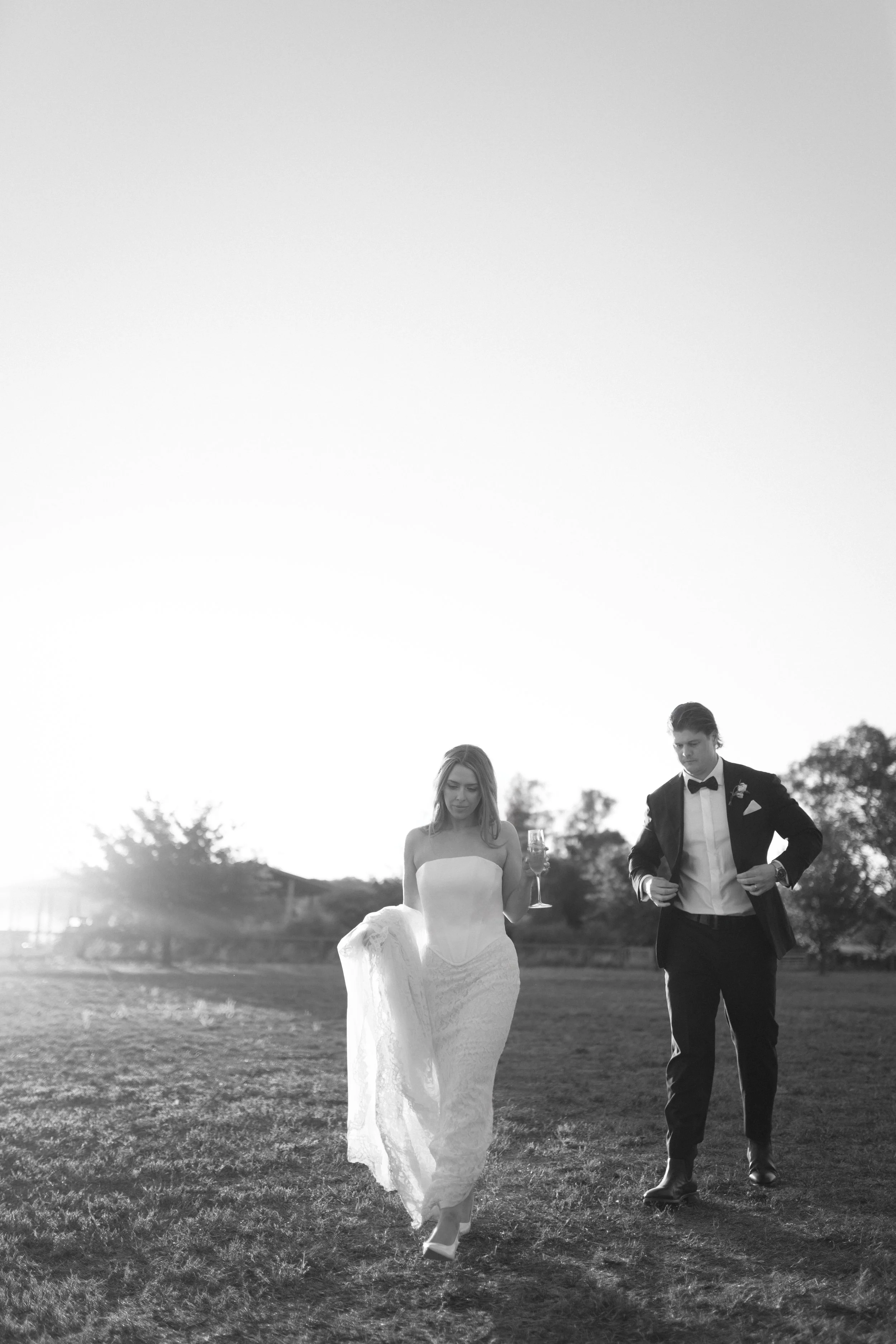 A couple dressed in formal wedding attire walking outdoors on grass, with trees and a setting sun in the background, in black and white.