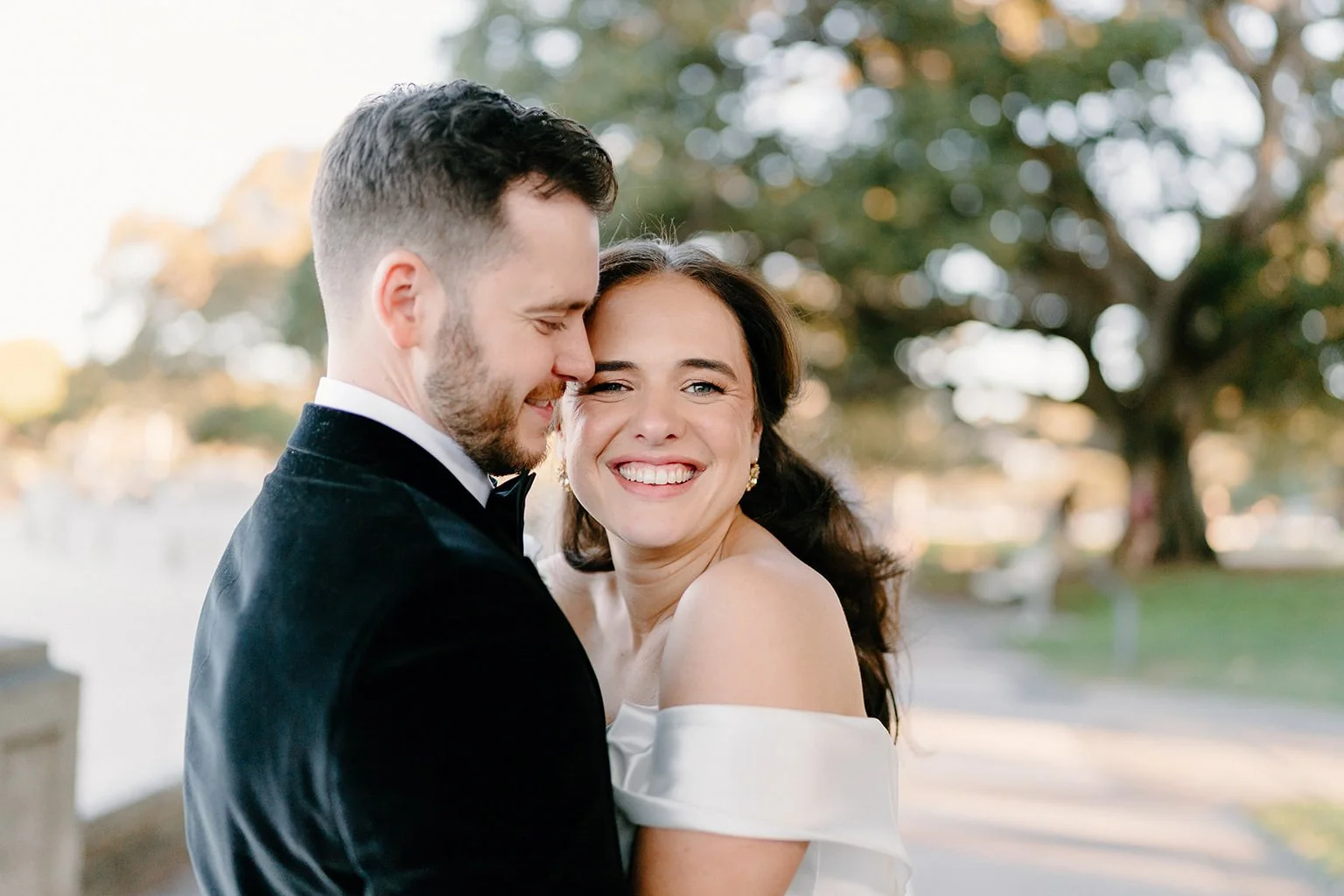 A smiling woman in an off-shoulder white dress and a beaming man in a tuxedo share a joyful moment outdoors with trees in the background.