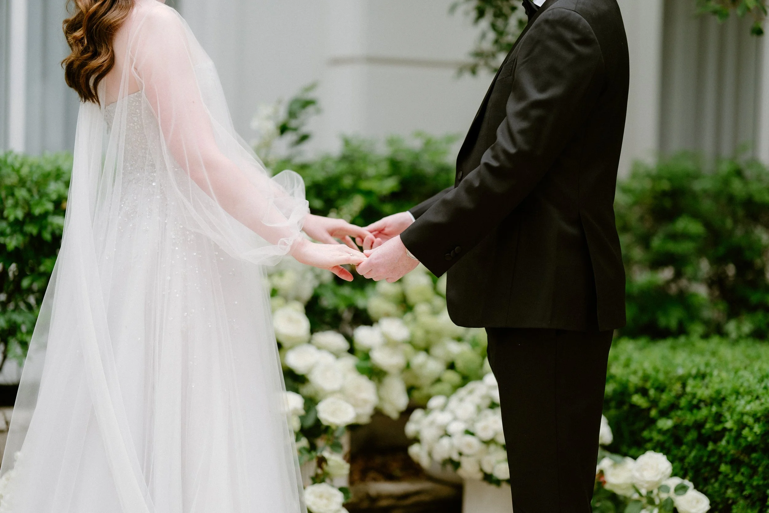A bride and groom holding hands during their wedding ceremony outdoors with white flowers and green bushes in the background.
