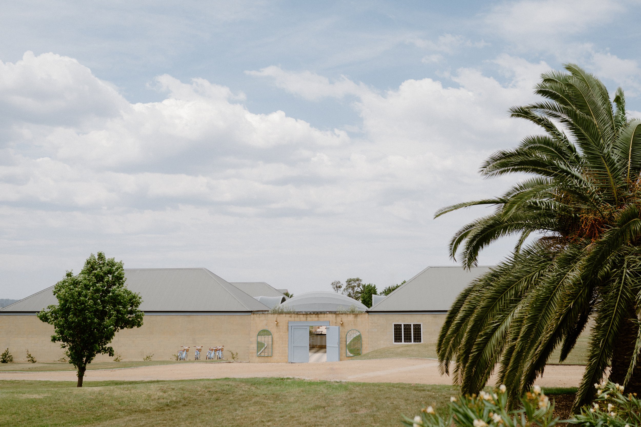 A large beige building with a metal roof, surrounded by a gravel pathway and greenery, including a tall palm tree and a smaller tree, under a partly cloudy sky.