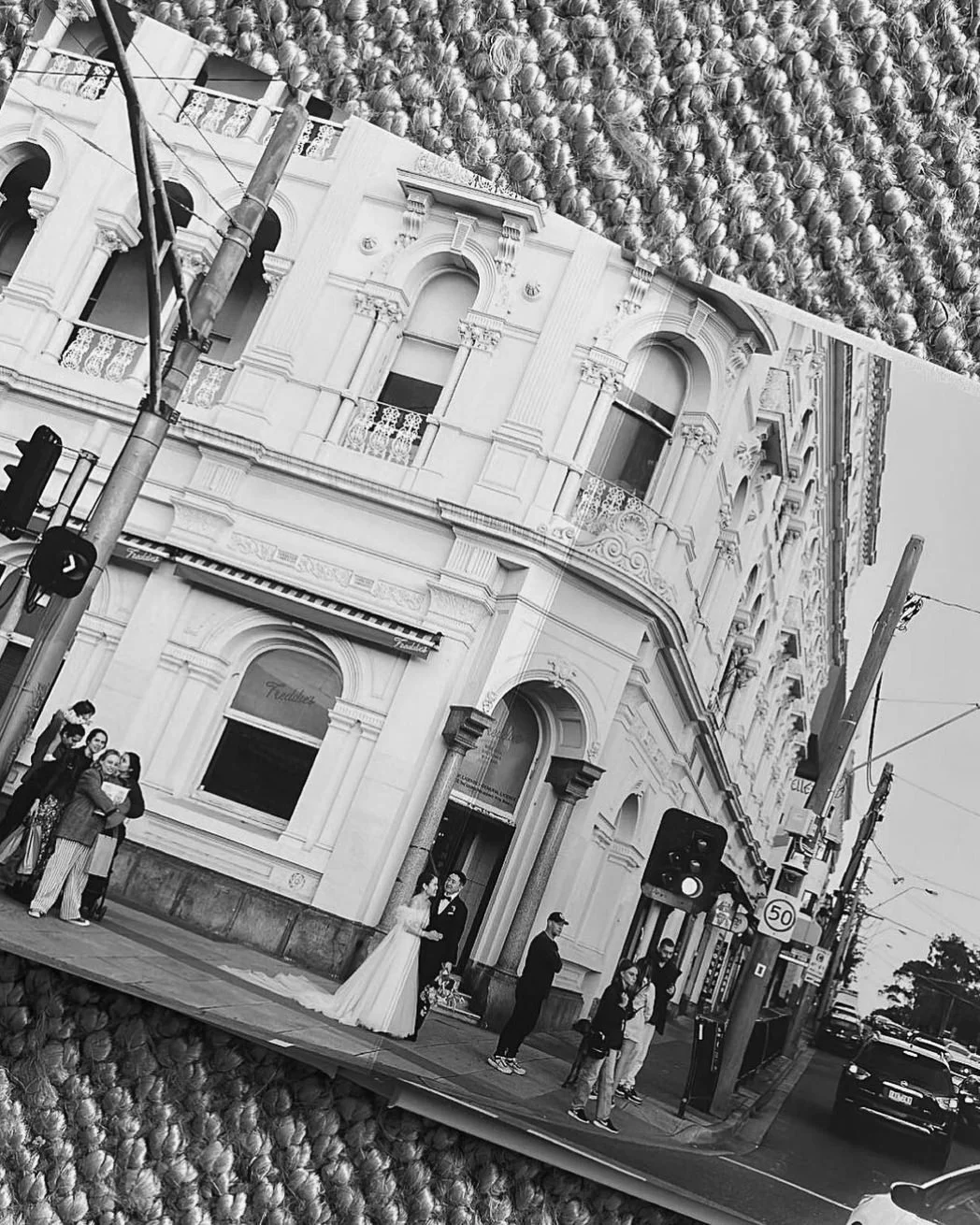Black and white photo of a city street with a historic building and a group of people, including a bride and groom, near the building's entrance.