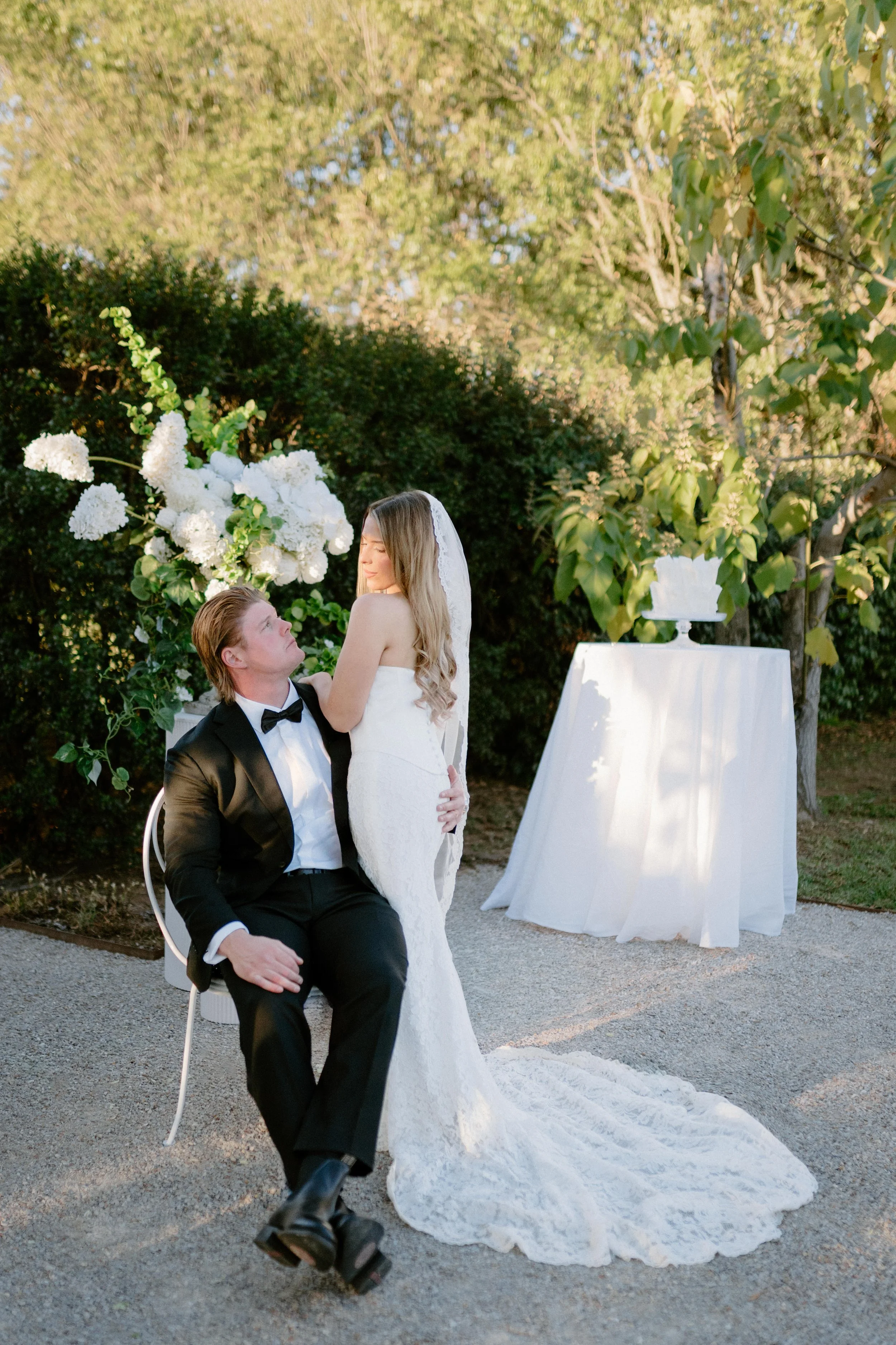 A bride and groom during an outdoor wedding ceremony. The groom is seated on a chair, wearing a black tuxedo with a bow tie, looking up at the bride. The bride, in a white wedding gown and veil, stands close to the groom. Behind them is a floral arra