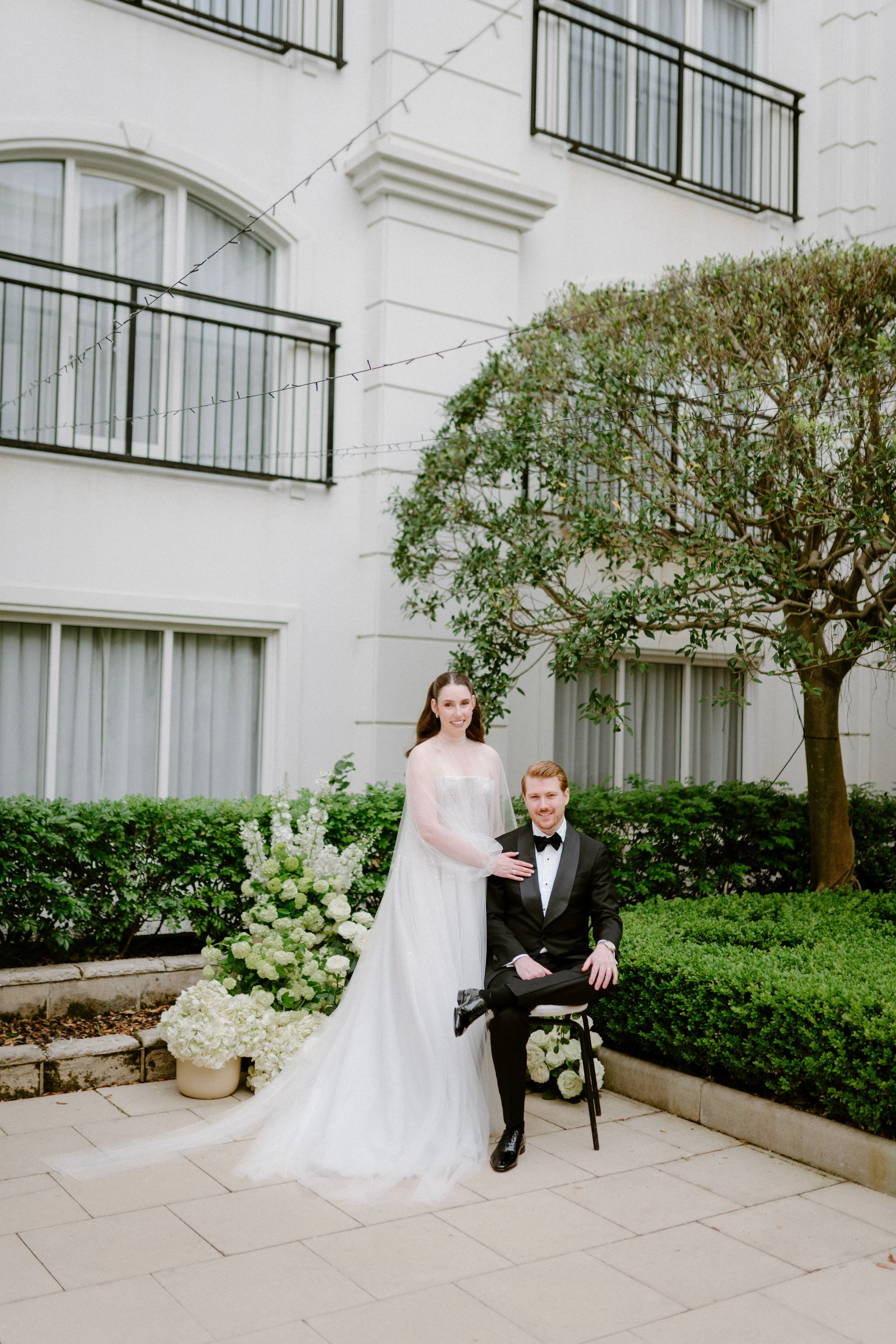 A bride in a white wedding gown and a groom in a black tuxedo are posing outdoors in front of a white building with balconies and windows, surrounded by lush green bushes and a small tree; the bride is standing, and the groom is seated on a chair.