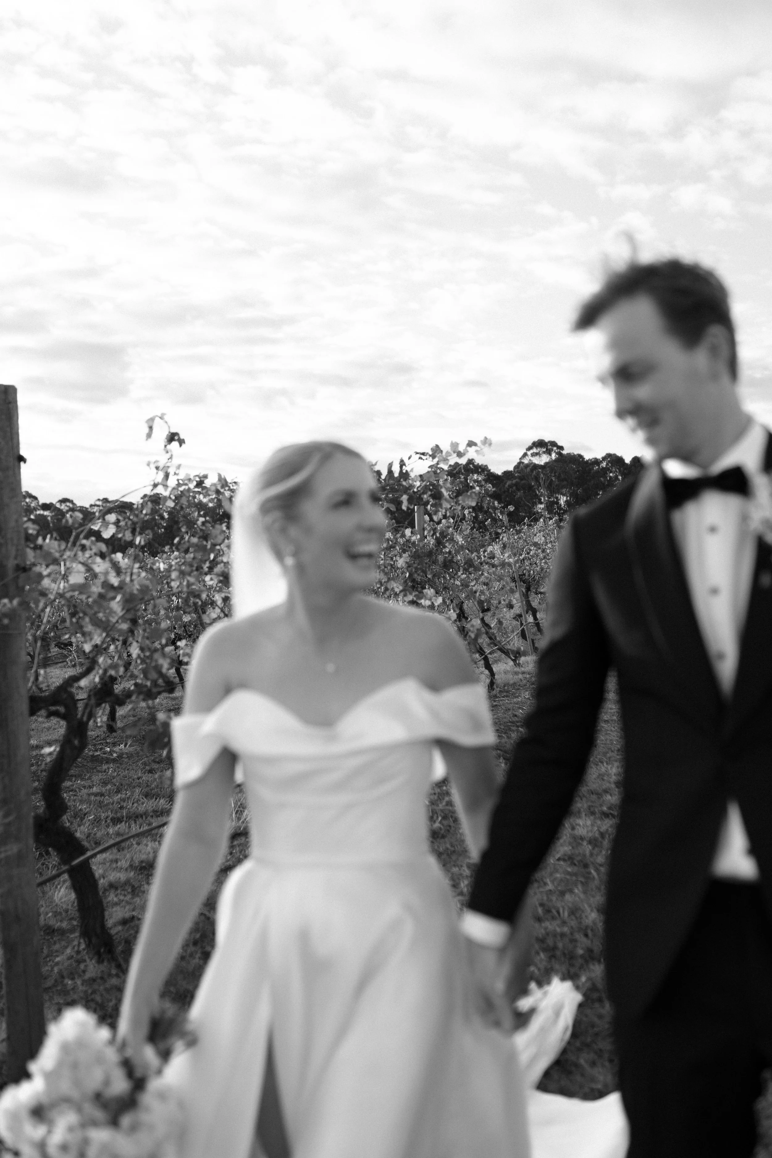 Black and white photo of a bride and groom holding hands in a vineyard, with the bride smiling and the groom looking at her, under a cloudy sky.