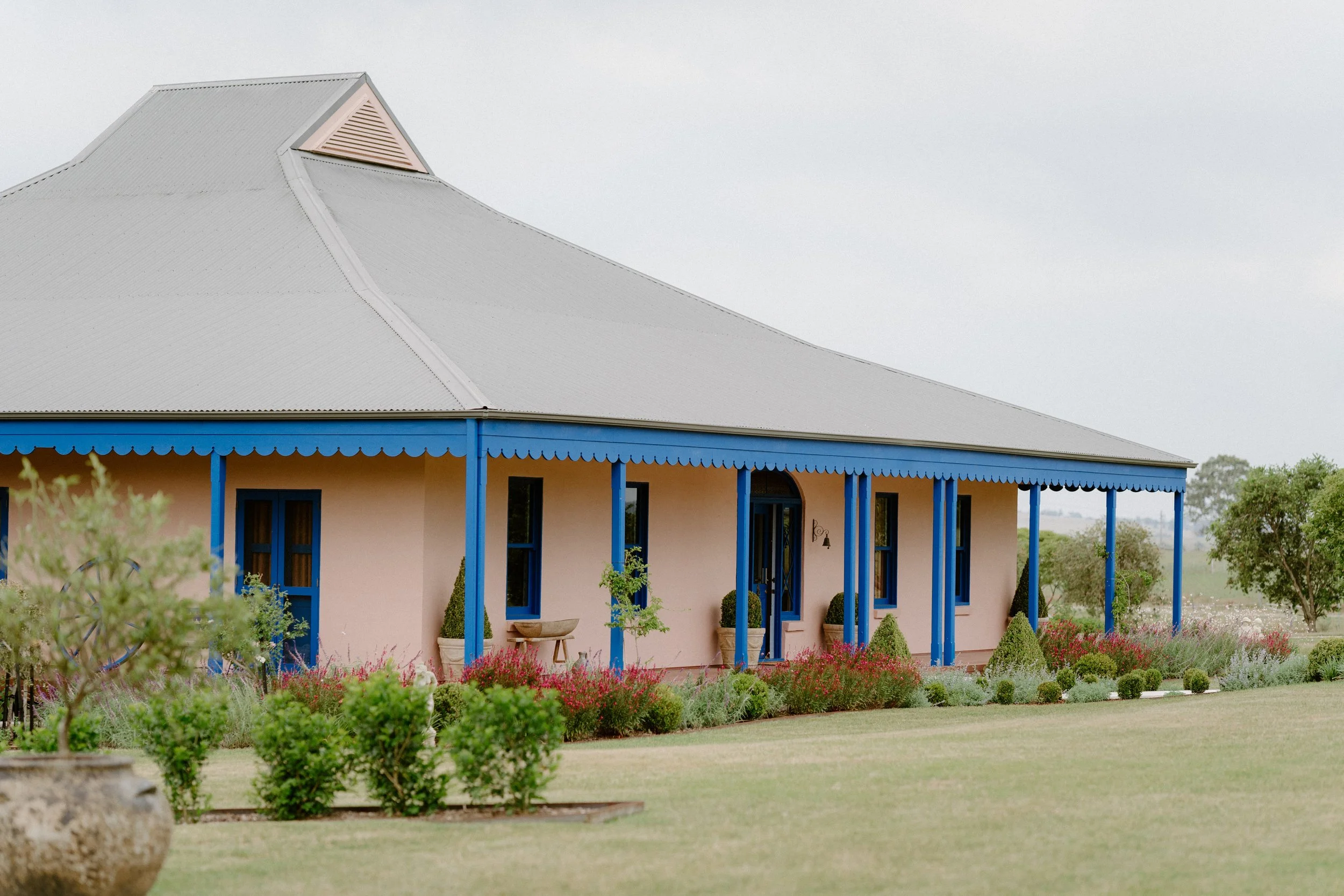 The Sienna, Hunter Valley Wedding Venue, Wedding Photos. A pink house with blue trim, metal roof, and a porch with blue columns. Surrounded by a garden with bushes and flowers.