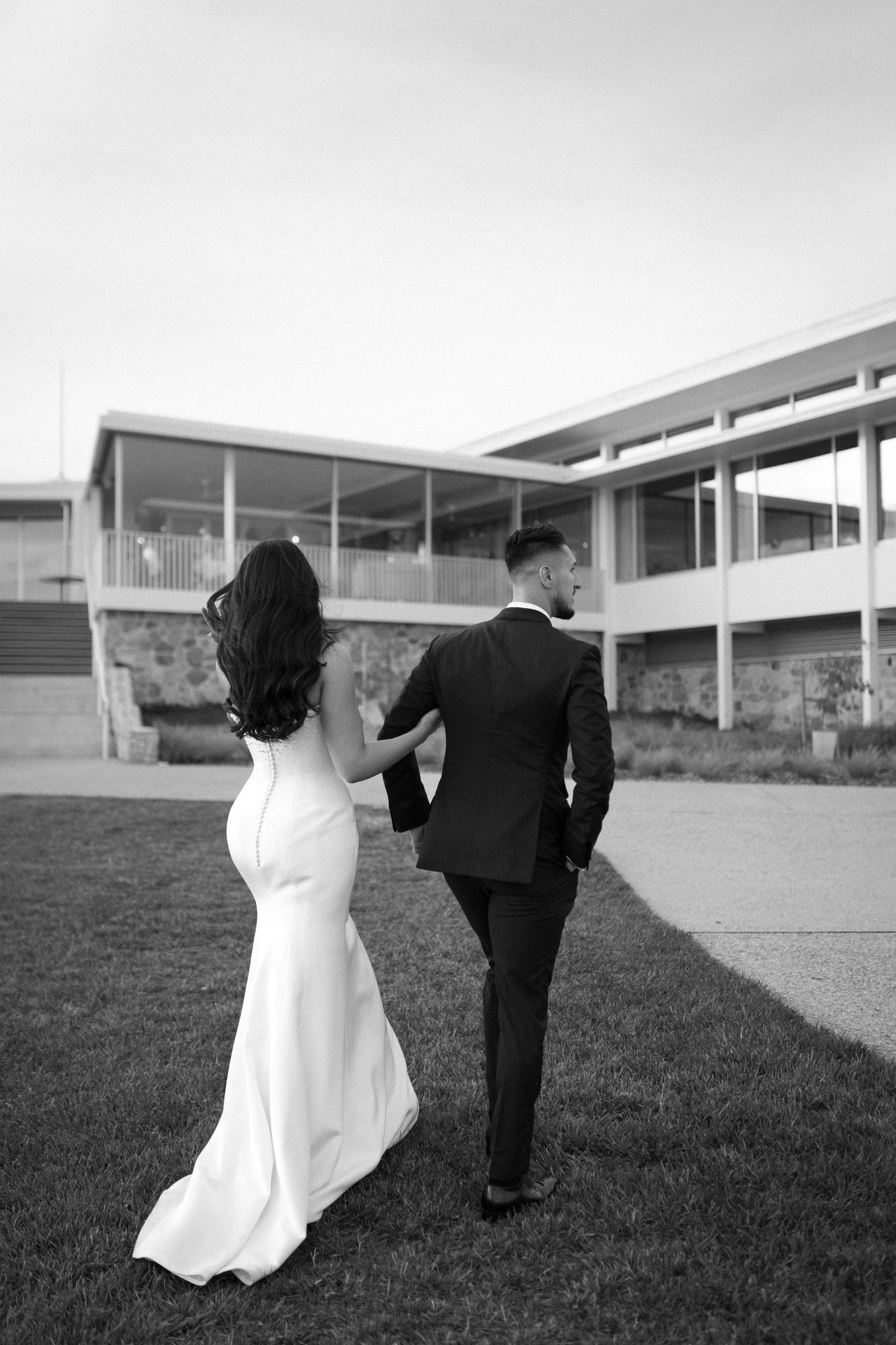A black and white photo of a bride and groom walking hand in hand on a lawn outside a modern building, with the bride in a white gown and the groom in a dark suit.