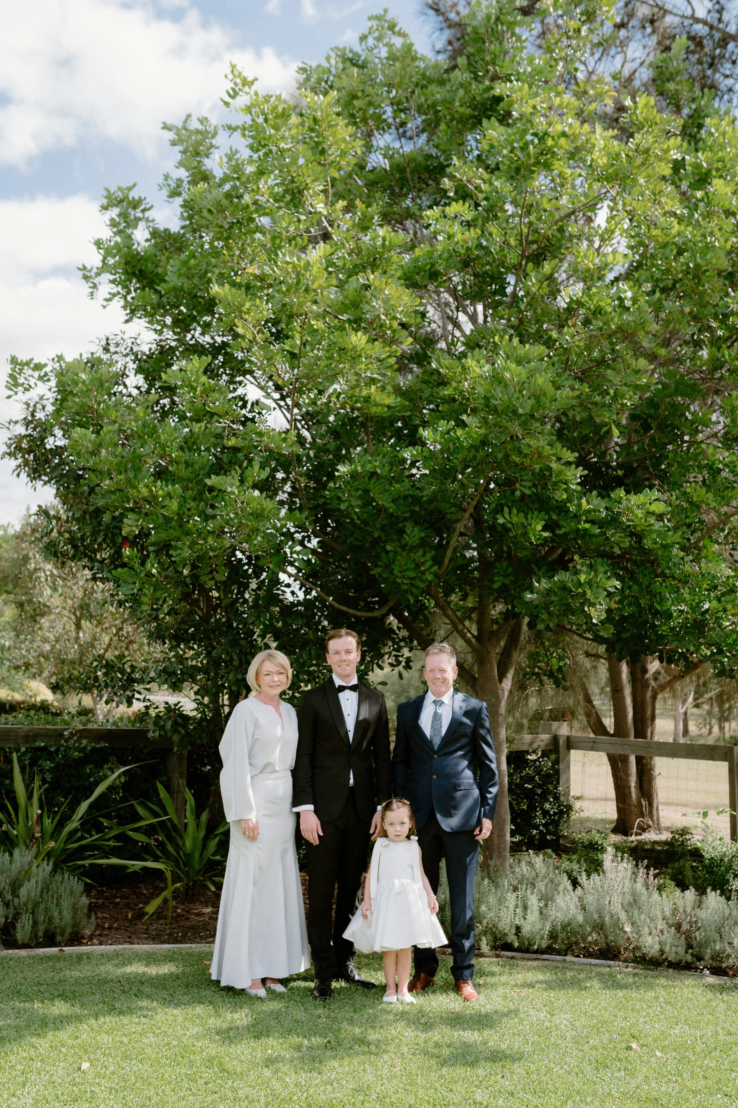 A family of five people dressed in formal attire, standing outdoors under a large tree with green leaves, on a sunny day with partly cloudy sky, in a garden or park setting.
