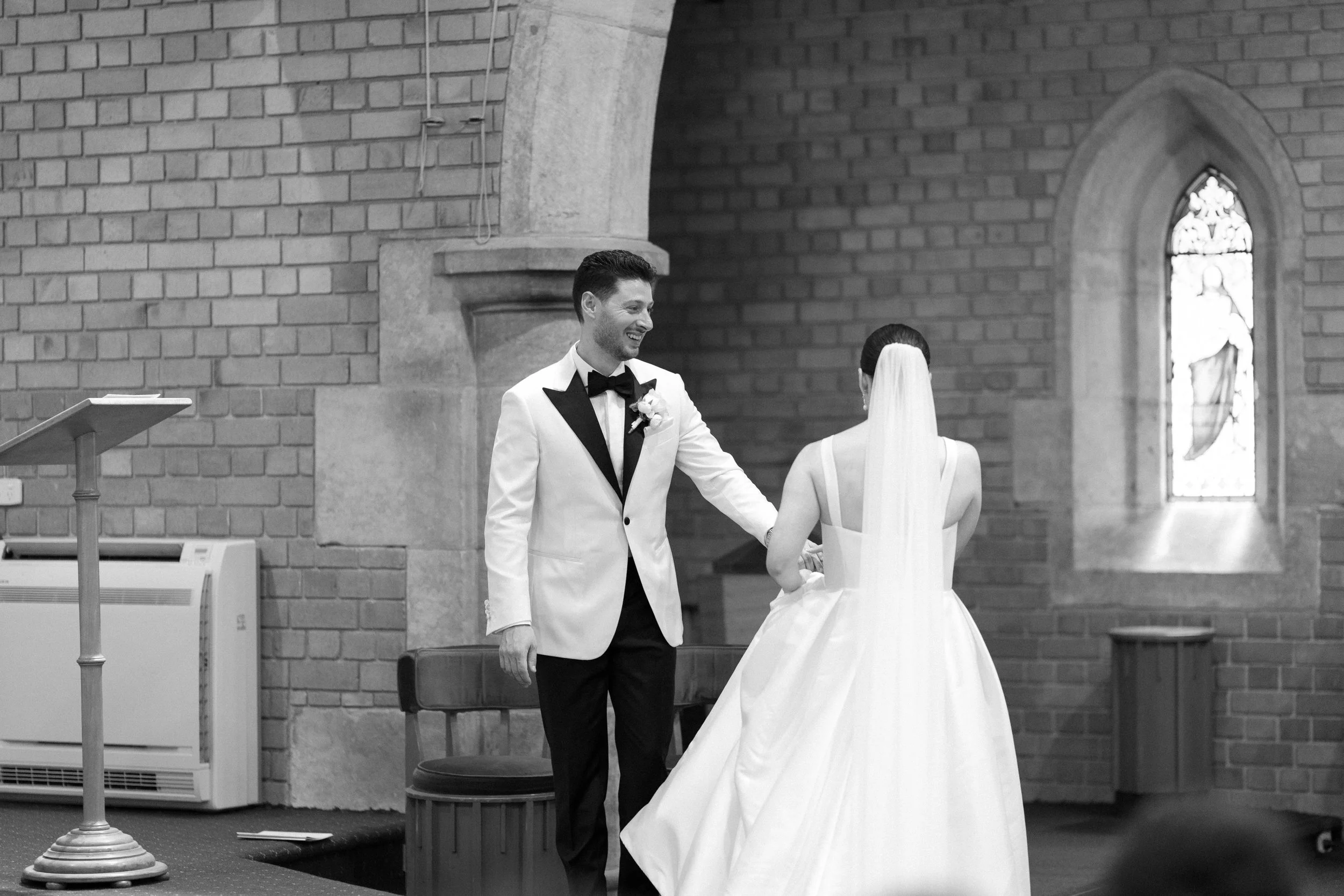 A groom in a white tuxedo with black lapels, smiling and holding hands with a bride in a white wedding gown with a veil, inside a brick church during a wedding ceremony.