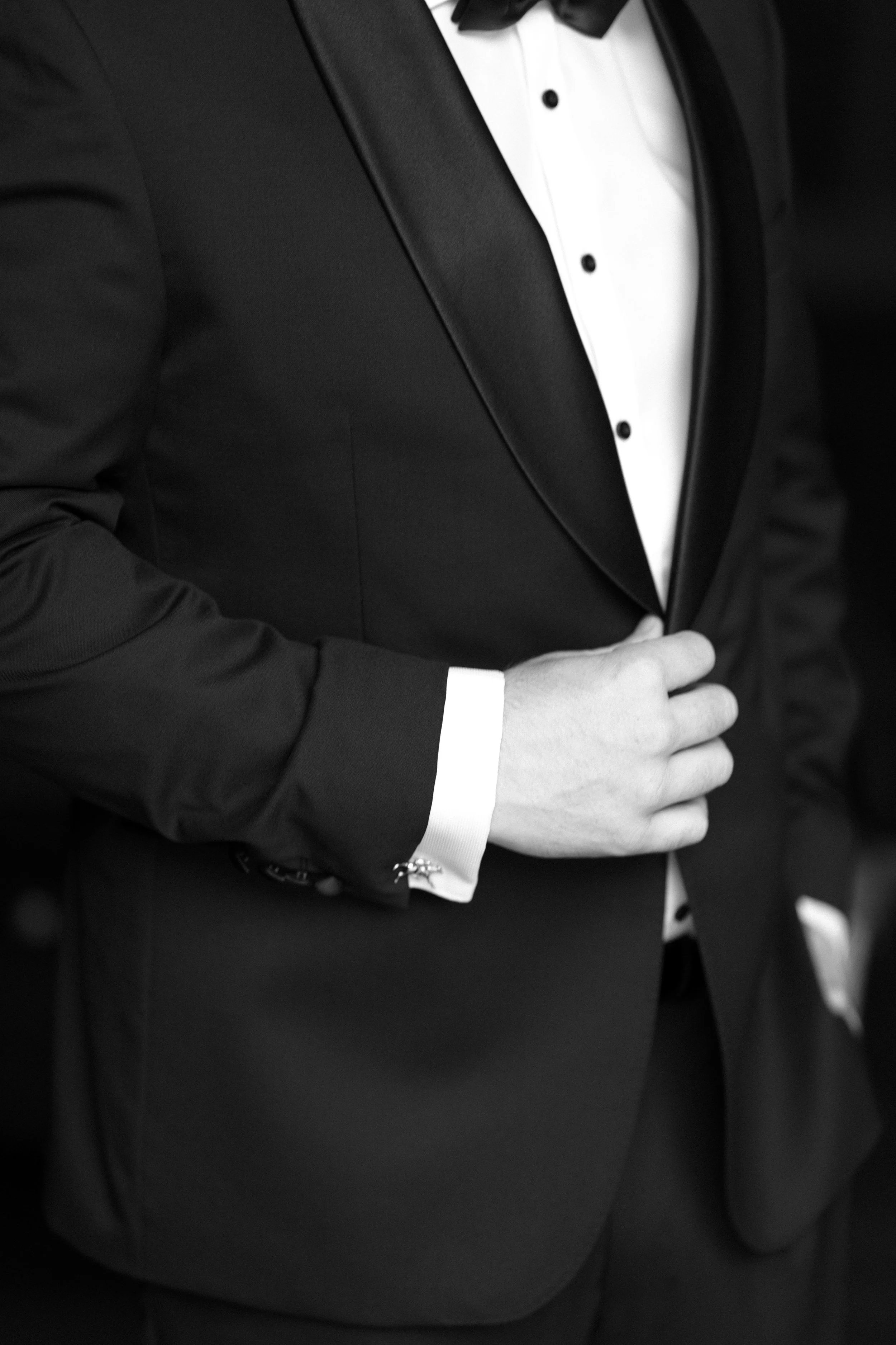 Close-up of a man in a black tuxedo adjusting his jacket, with a white dress shirt and black bow tie, black and white photograph.
