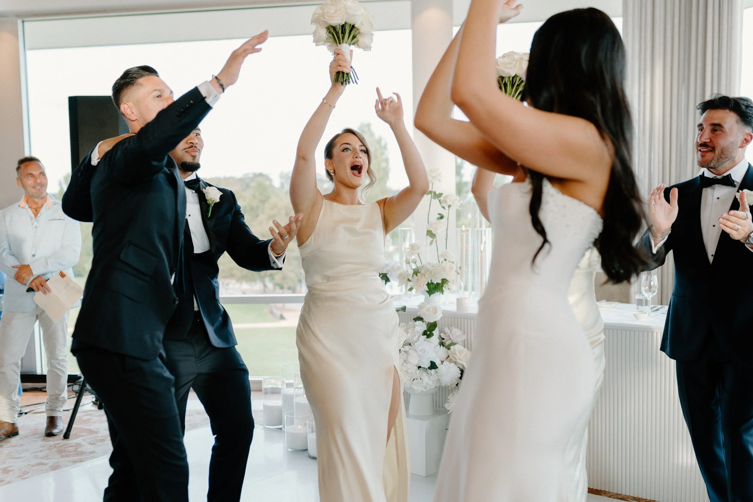 People dancing and celebrating at a wedding reception, with women in white dresses and men in tuxedos, holding bouquets and clapping