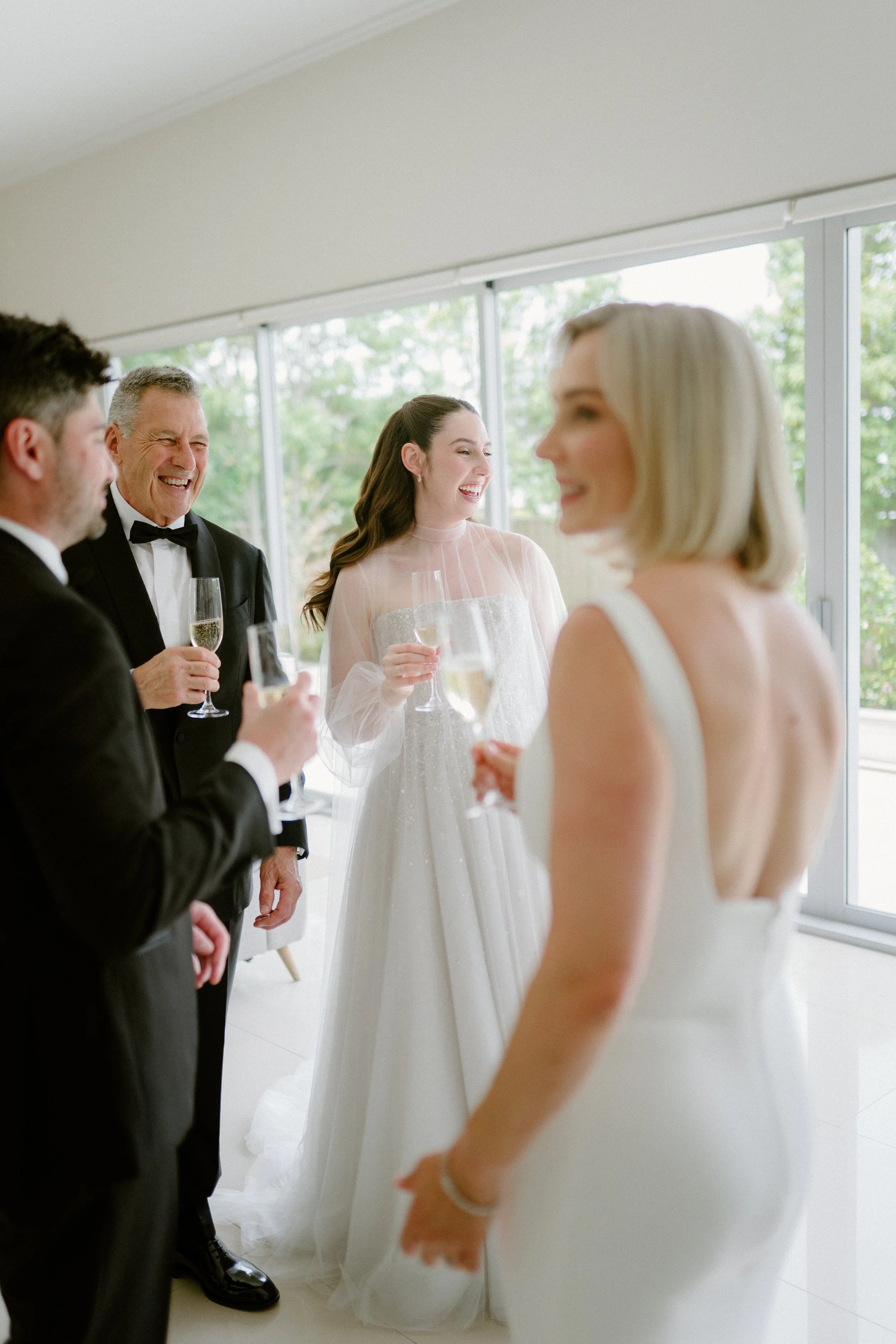 People in formal attire celebrating with glasses of champagne on a bright indoor patio.