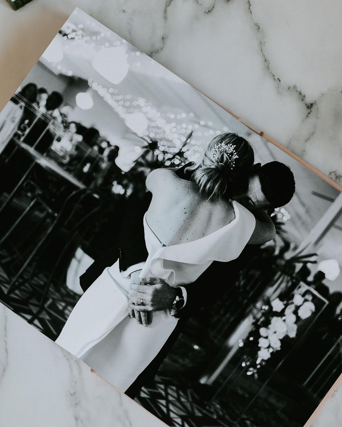 A black and white photo of a couple hugging at their wedding reception, with guests seated at tables and decorative string lights in the background.