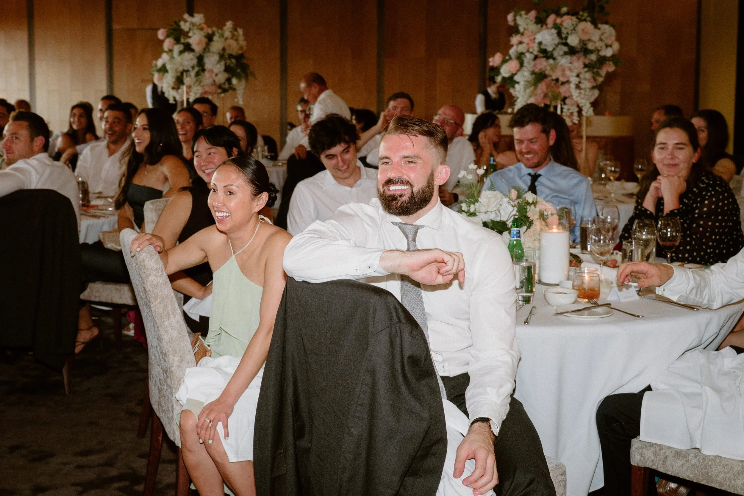 People sitting at a round table during a wedding reception, smiling and laughing, with floral centerpieces and candles on the table.