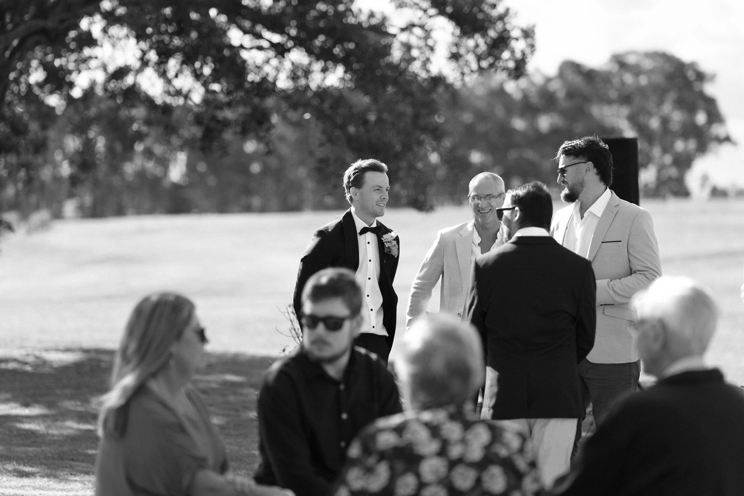 A black-and-white photo of a wedding ceremony outdoors, with the groom in a tuxedo smiling and people sitting and standing around, some wearing sunglasses and formal attire, with trees in the background.
