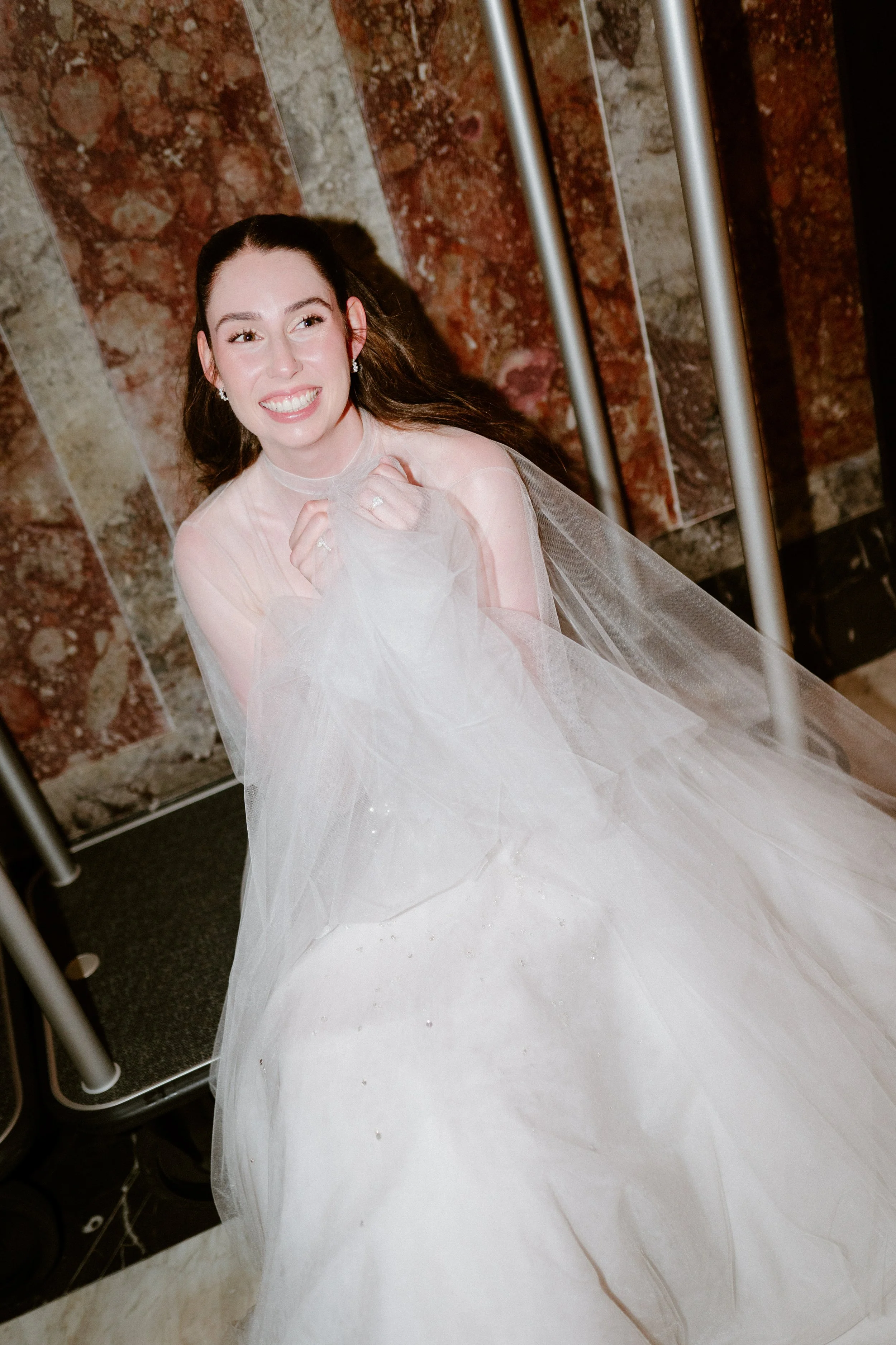 Smiling woman in a wedding dress sitting on a bench with a marble wall background.