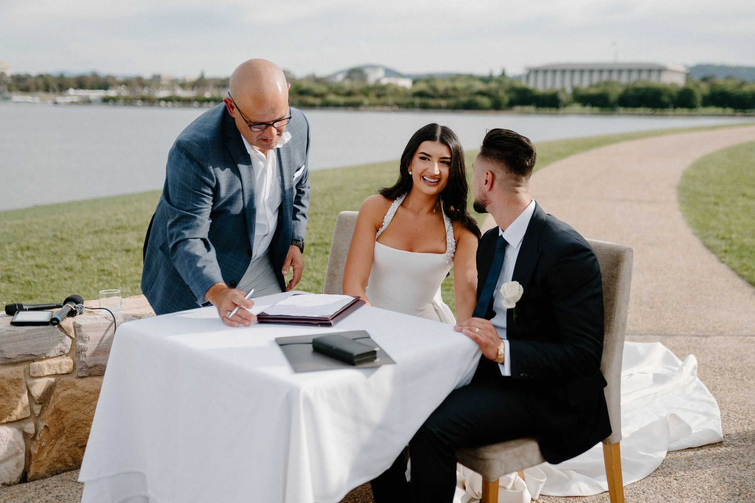 A couple getting married outdoors by the water, sitting at a table with a officiant signing a document, with a scenic background of a lake and green landscape.