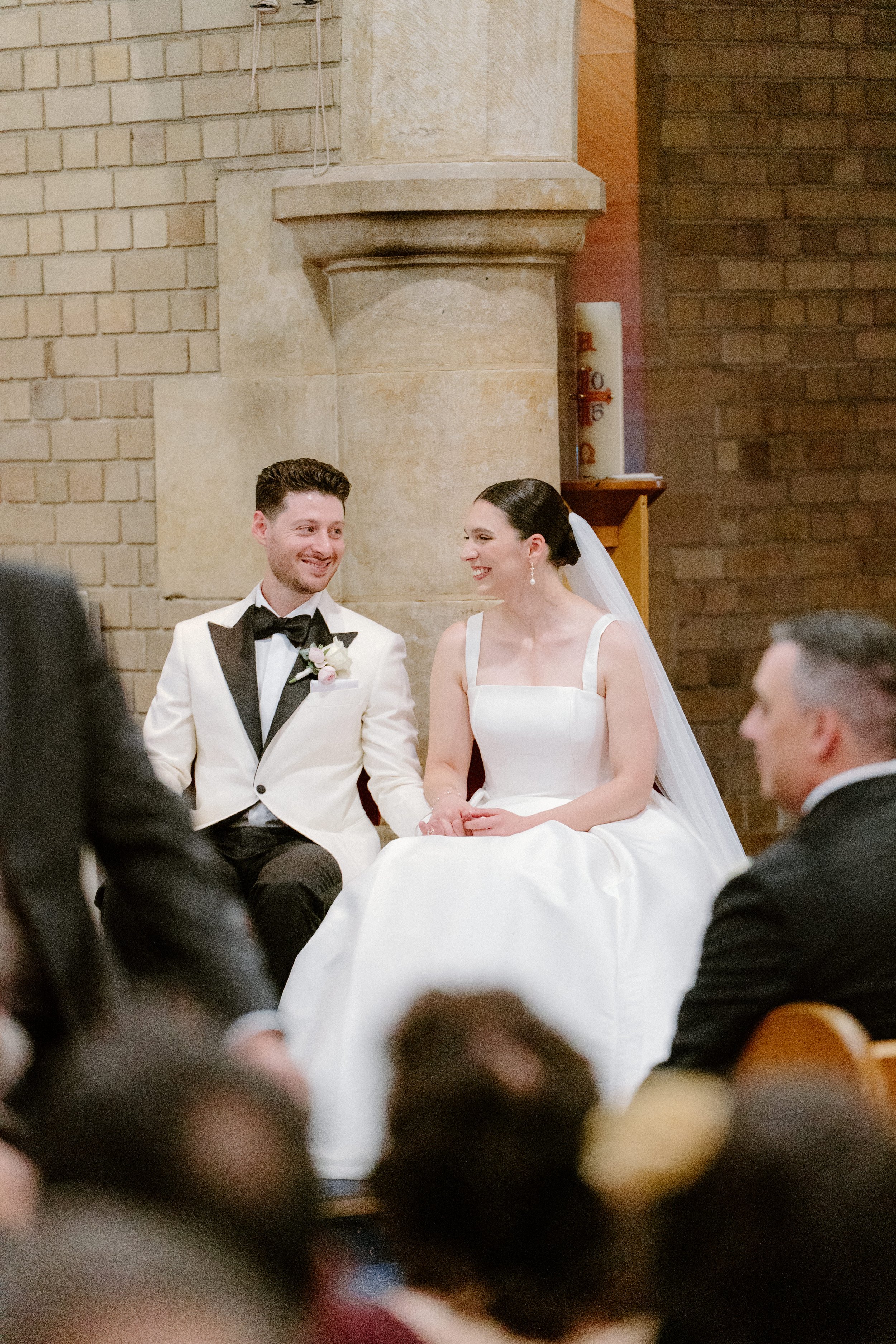 A bride and groom sitting together during their wedding ceremony inside a church, smiling at each other.